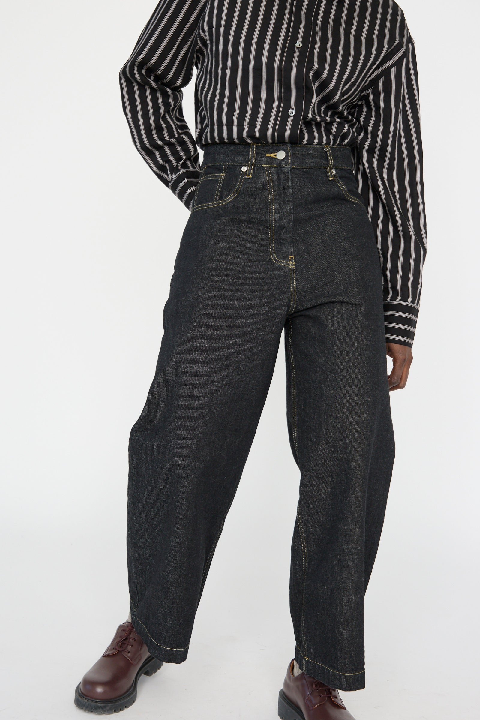 Person wearing a black and white striped shirt, Italian Denim Neive Pant in Black by Studio Nicholson, and dark brown shoes, one arm by their side against a white background.