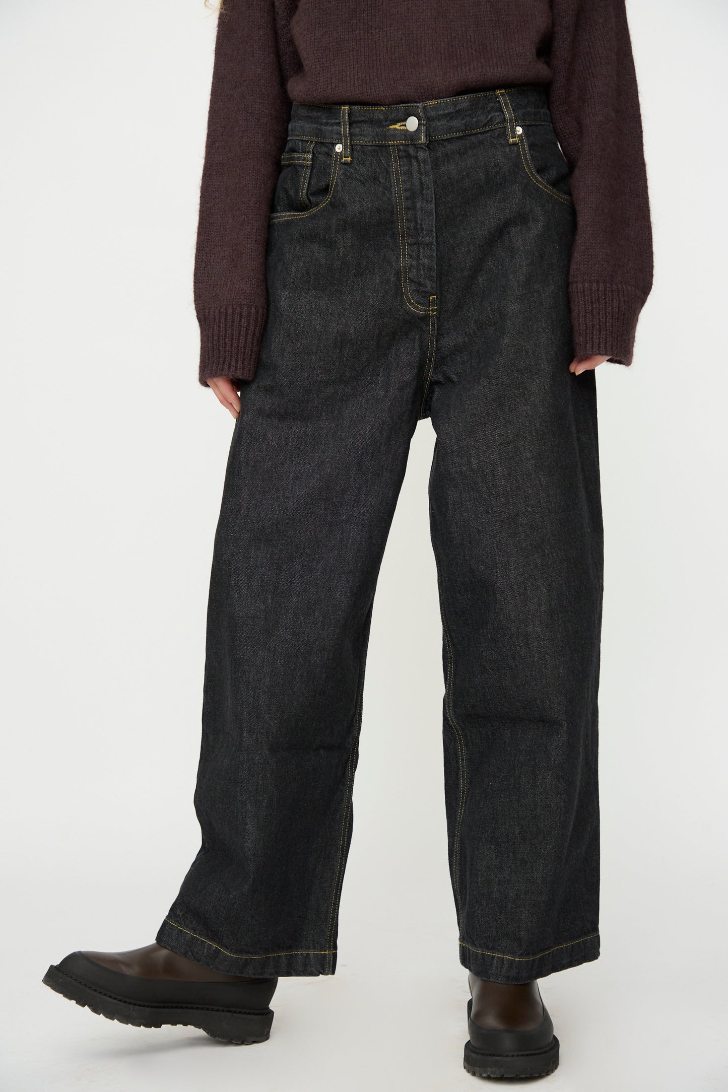 A person wears Studio Nicholson's Italian Denim Neive Pant in Black, styled with a dark long-sleeve sweater and black boots, standing against a plain light background.