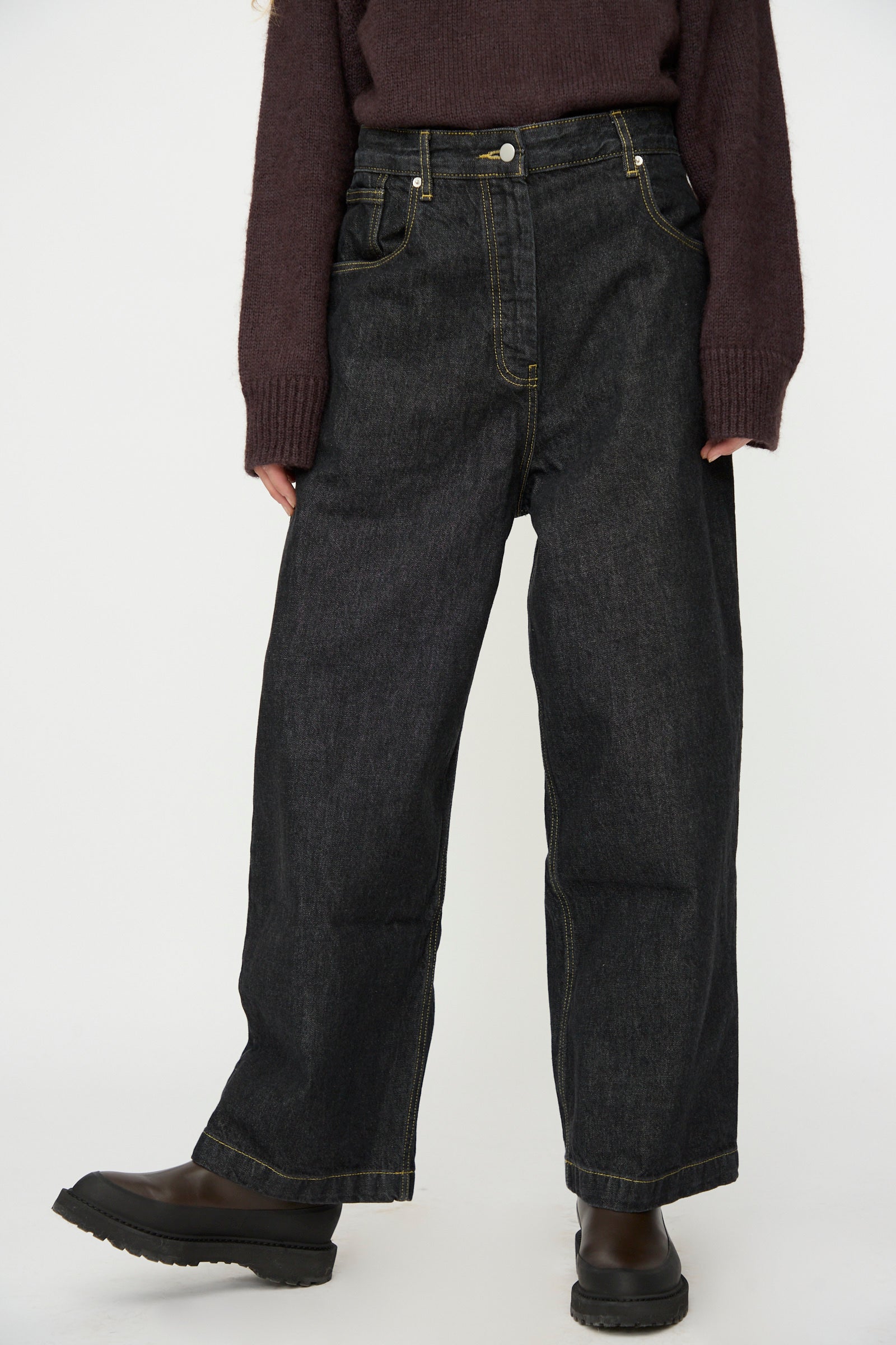 A person wears Studio Nicholson's Italian Denim Neive Pant in Black, styled with a dark long-sleeve sweater and black boots, standing against a plain light background.
