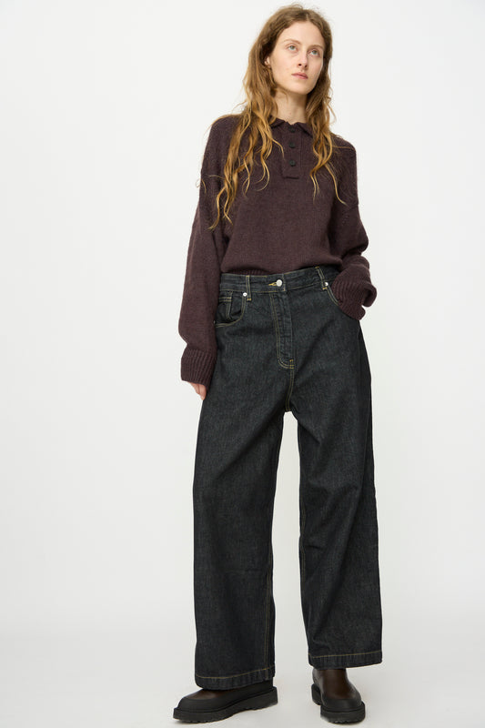 A person with long, wavy hair wears a dark purple sweater, Studio Nicholson’s Italian Denim Neive Pant in Black, and black shoes while standing against a plain white background.