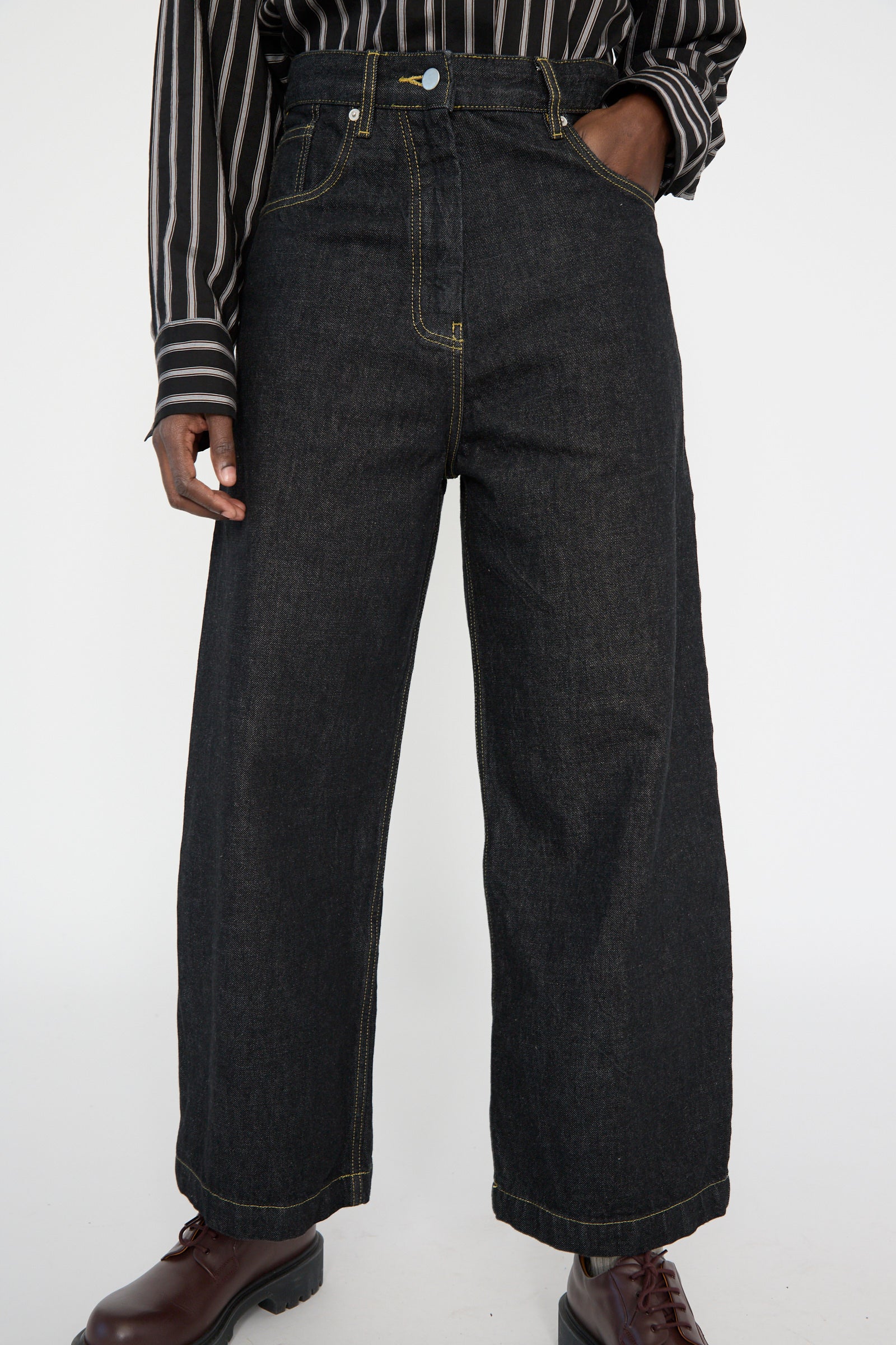 A person wears Studio Nicholson's Italian Denim Neive Pant in Black, expertly paired with a striped shirt and brown boots against a plain background.