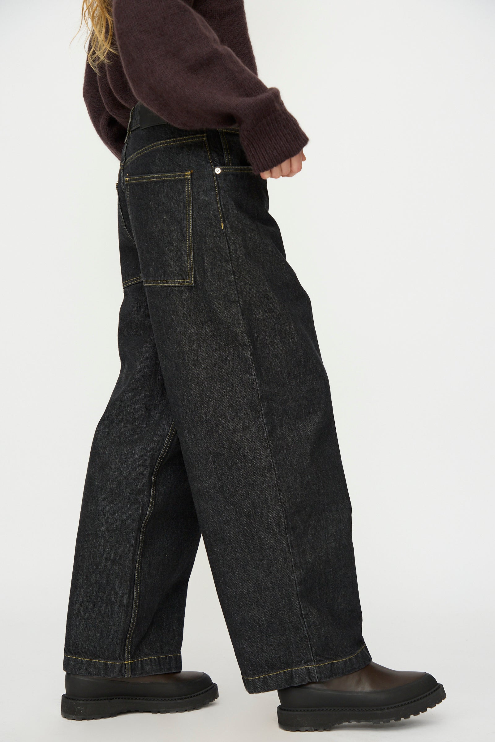 A person, partially visible from the shoulders down, wears Studio Nicholson's Italian Denim Neive Pant in Black, paired with a brown sweater and black shoes, standing against a plain white background.