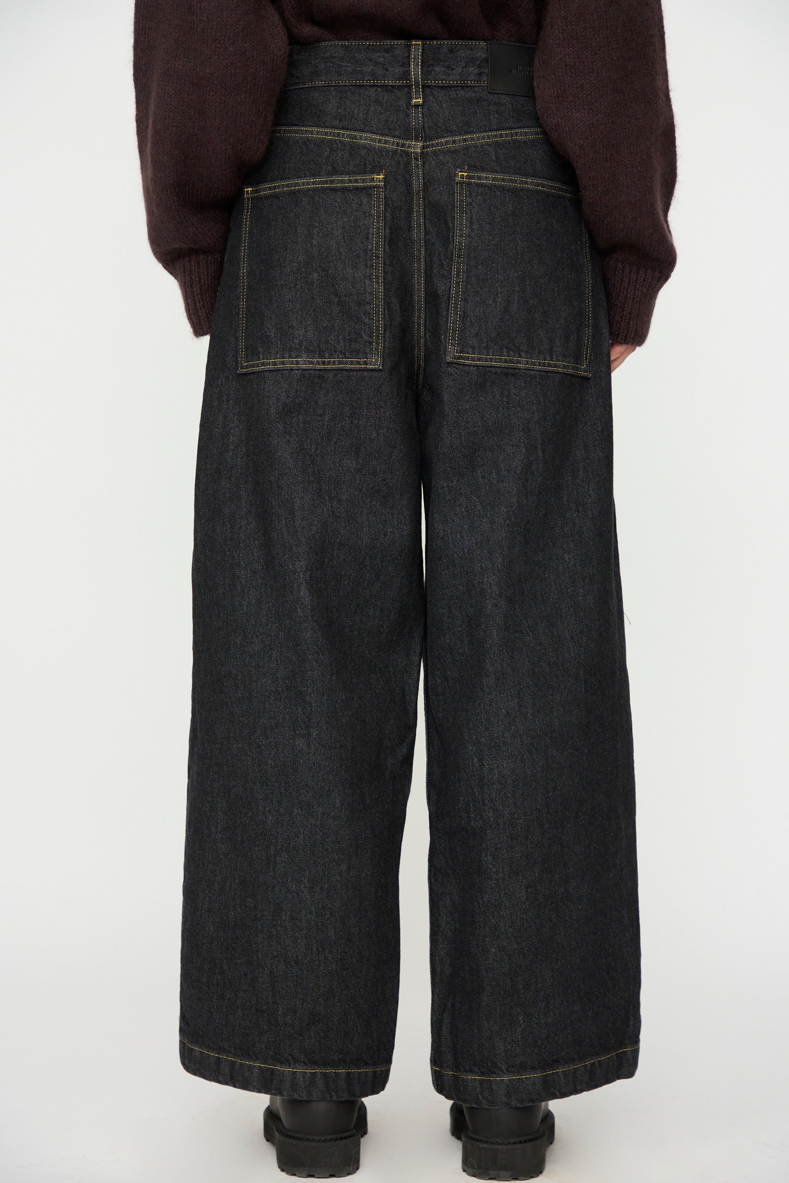 Model wears Studio Nicholson's Italian Denim Neive Pant in Black, featuring wide legs and large back pockets, paired with a dark sweater. Shown from the back against a plain light background.