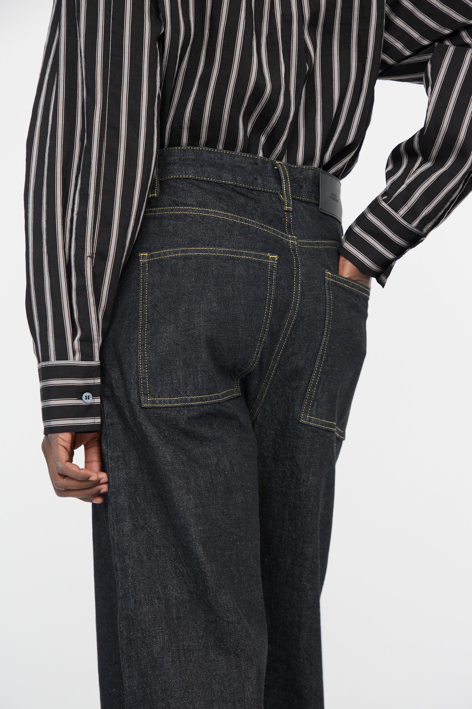 Side view of a person in a black and white striped shirt, wearing Studio Nicholson's Italian Denim Neive Pant in Black with distinctive yellow stitching, hands in back pockets.