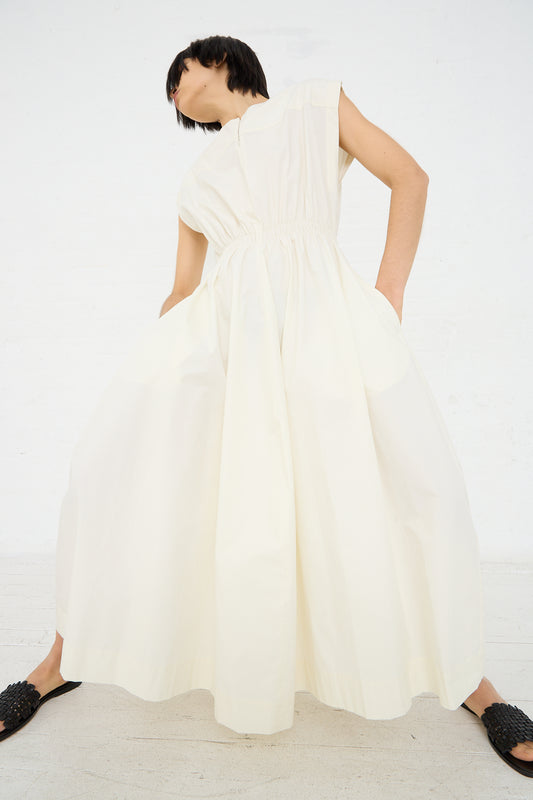 A woman stands with her back to the camera, wearing a Studio Nicholson Katrine Ruched Waist Dress in Parchment that reaches the floor, with her head turned to one side, set against a plain white background.