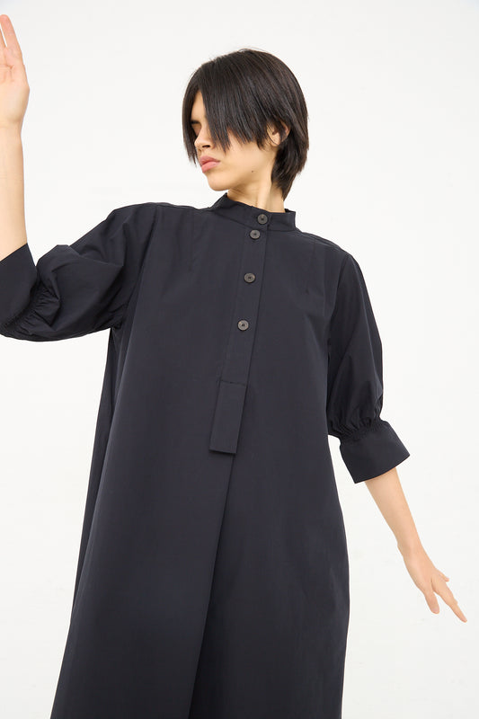 A person with short hair wearing a Studio Nicholson Knoll Shirt Dress with Ruched Cuff in Darkest Navy poses with one arm raised and the other extended downwards against a white background.
