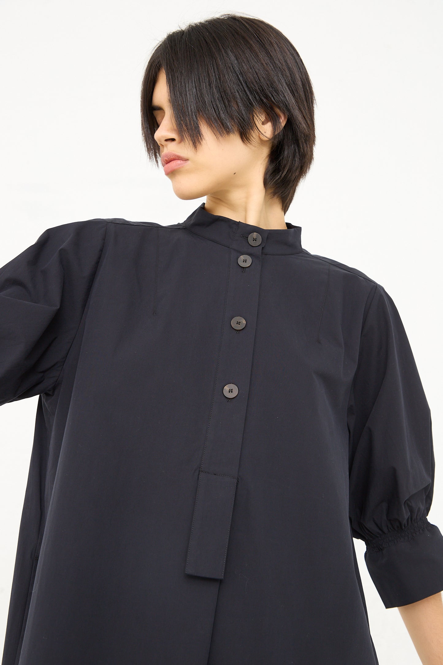 A woman with a short bob haircut is wearing a dark navy Knoll shirt dress with ruched cuff by Studio Nicholson, looking to the side against a white background.