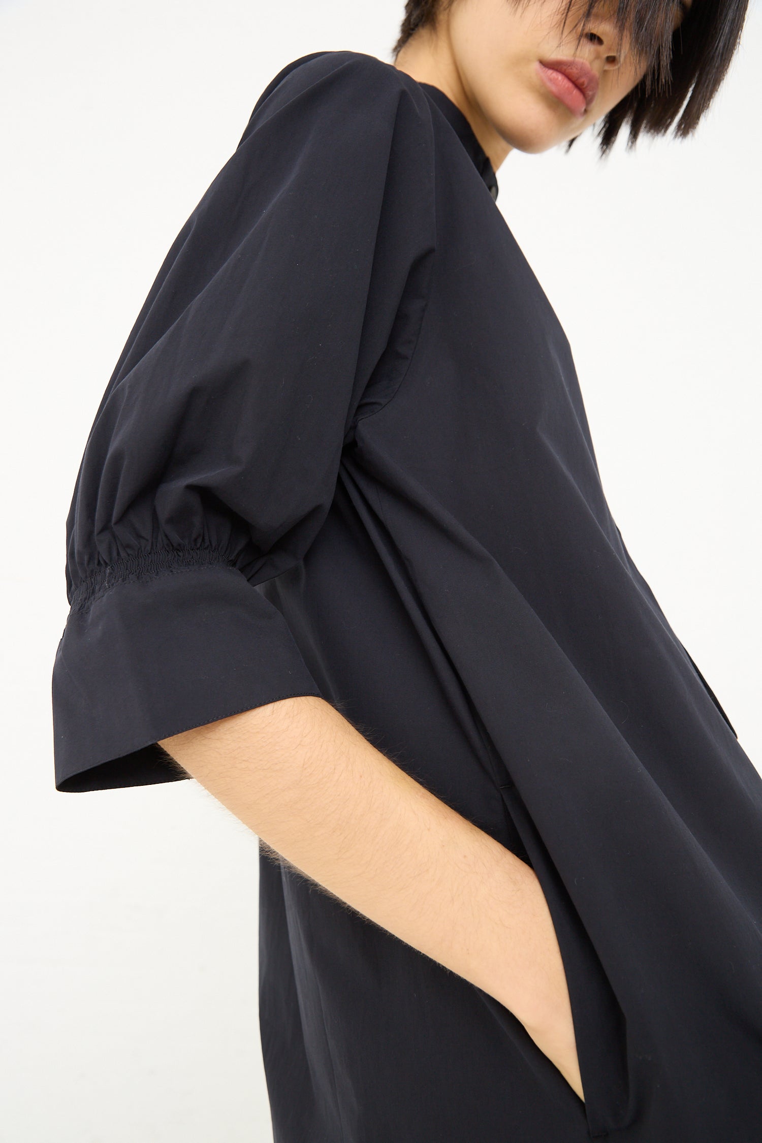 A person wearing a Knoll Shirt Dress with Ruched Cuff in Darkest Navy by Studio Nicholson stands against a white background, partially facing the camera.