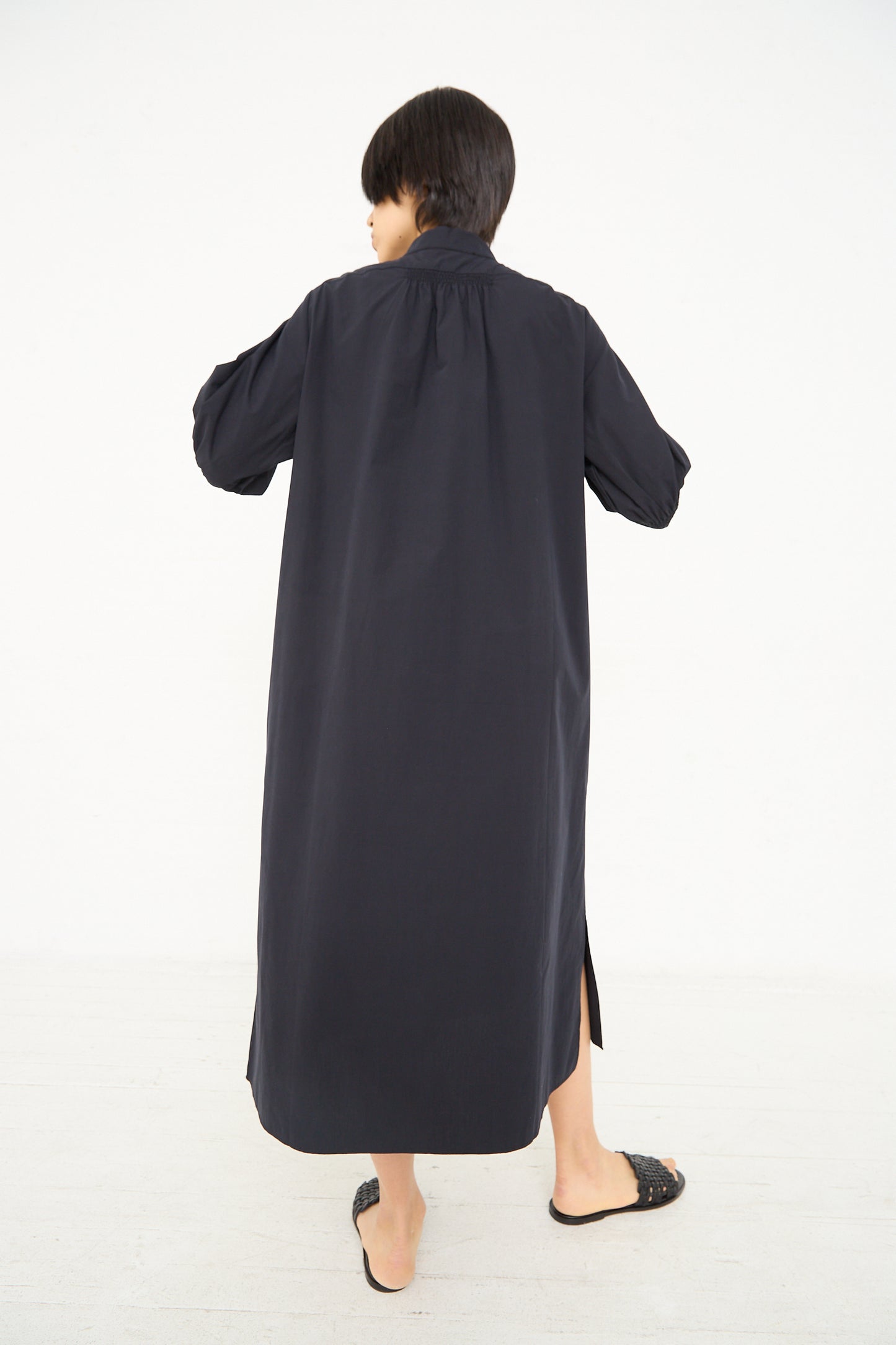 A person seen from behind wearing a Knoll Shirt Dress with Ruched Cuff in Darkest Navy by Studio Nicholson with puffed sleeves and black flats.