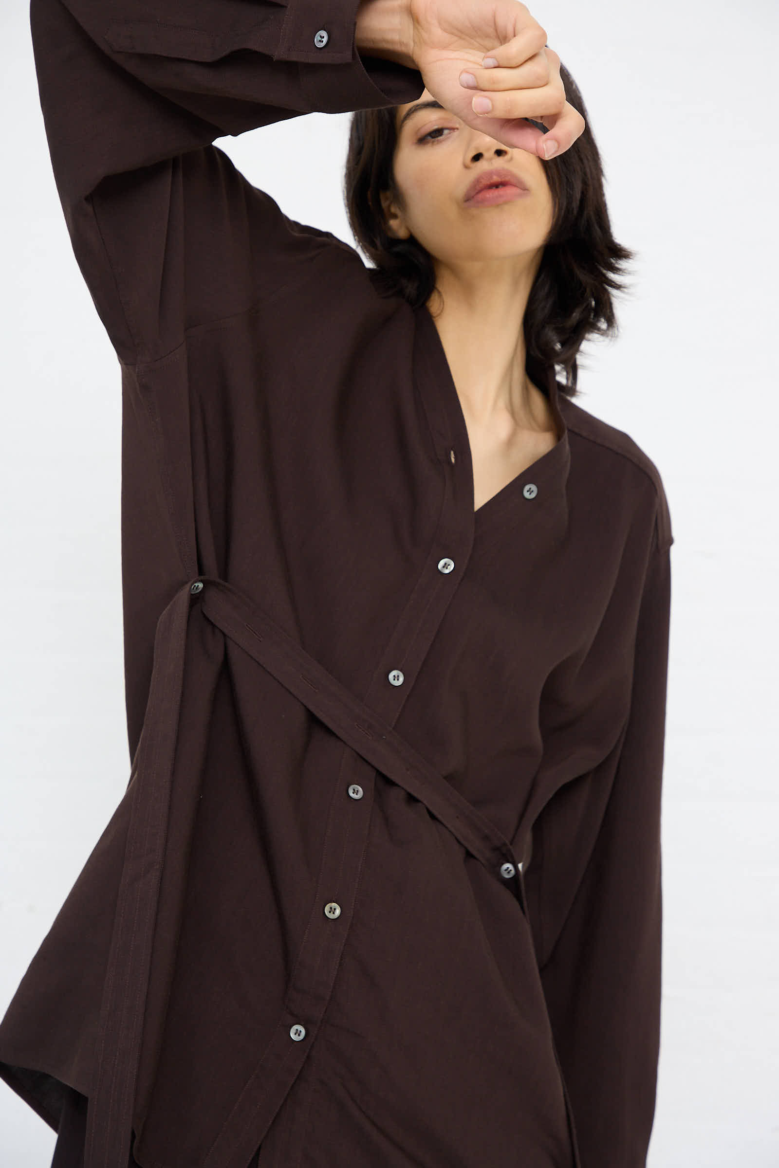 A person with short dark hair, wearing the Lane Collarless Shirt in Espresso from Studio Nicholson, poses with one arm raised in front of their face. Their expression is neutral, and the background is plain white.