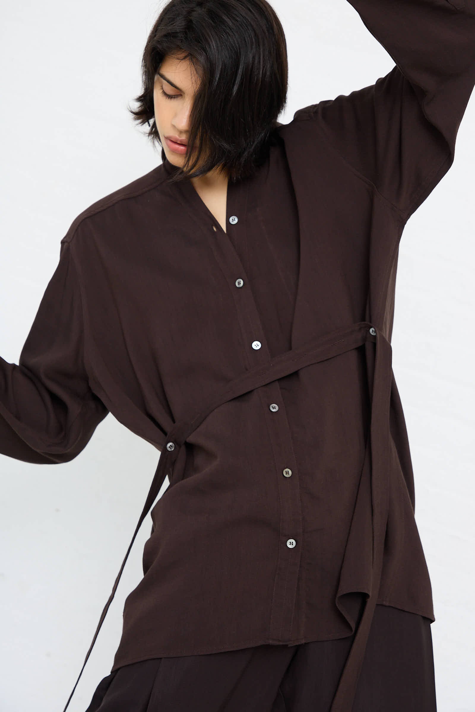 A person with shoulder-length dark hair wears the loose-fitting Lane Collarless Shirt in Espresso by Studio Nicholson, paired with black pants. They are standing against a plain white background with their head slightly tilted.