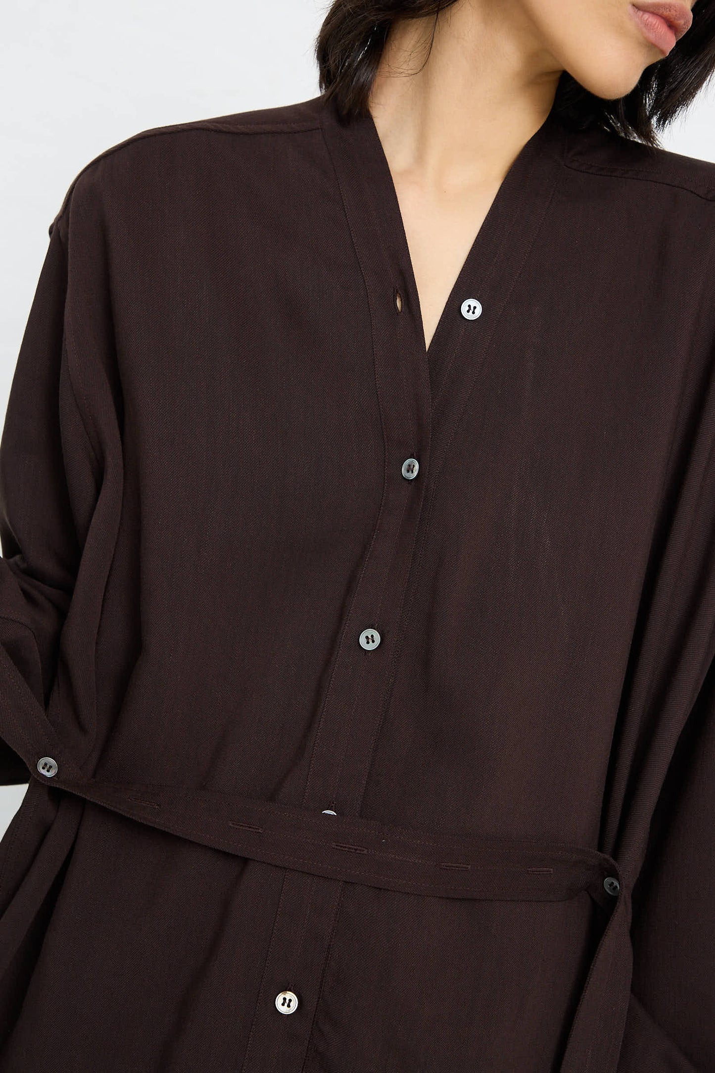 A person wearing the Lane Collarless Shirt in Espresso by Studio Nicholson, accessorized with a detachable belt tied at the waist. The shirt showcases a V-neck collar and long sleeves. The image is cropped to show only the upper part of their body.