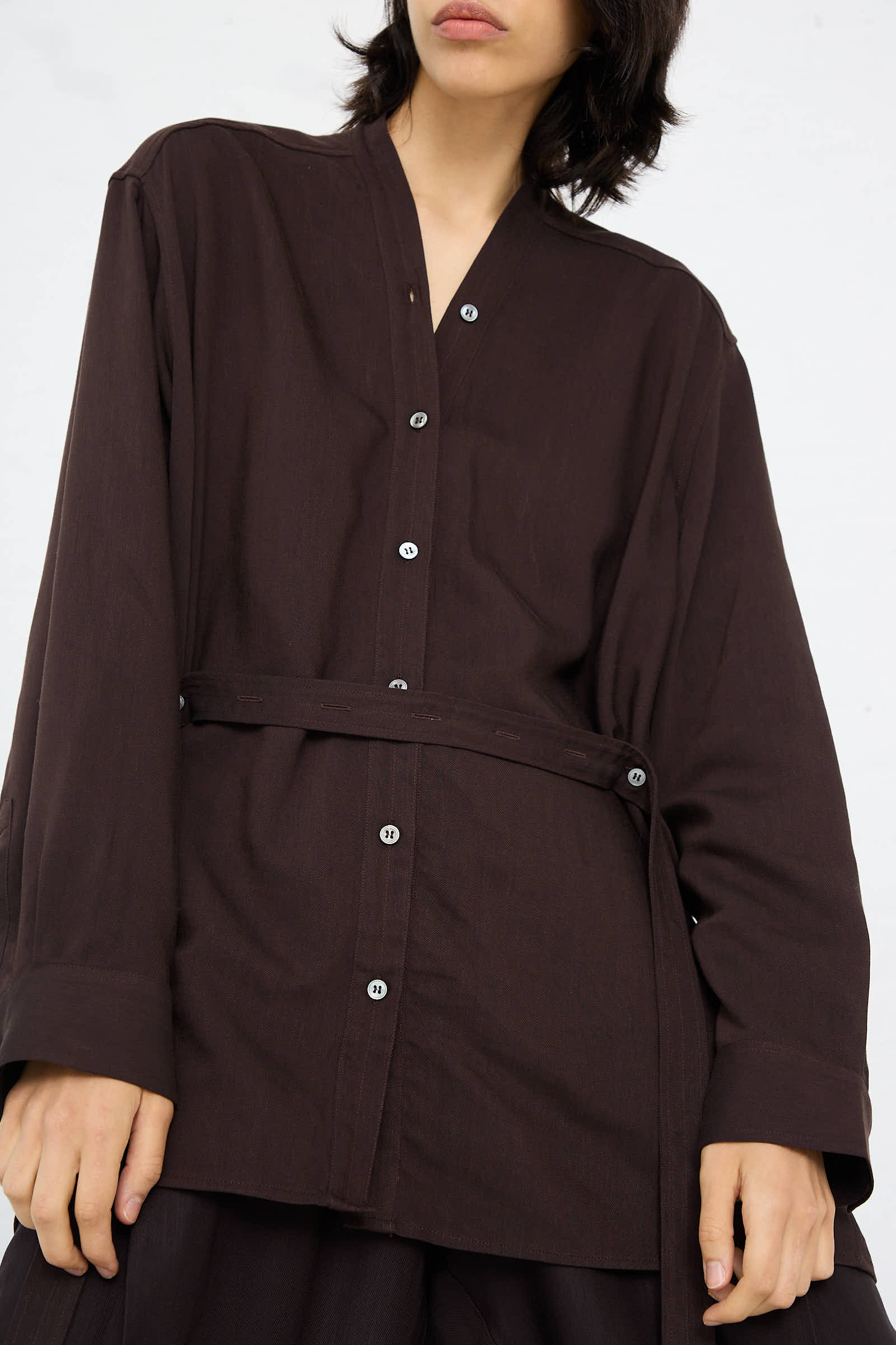 A person wearing the Studio Nicholson Lane Collarless Shirt in Espresso, crafted from soft viscose linen with long sleeves and a V-neck, stands against a plain background.