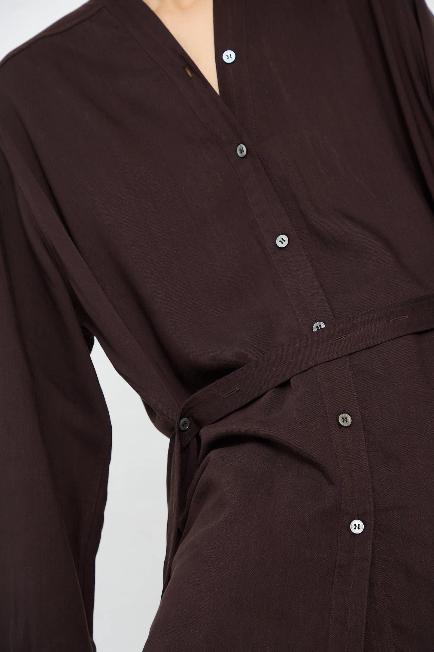 Close-up of a person wearing the Lane Collarless Shirt in Espresso by Studio Nicholson. Crafted from soft viscose linen, this dark brown shirt features a V-neck, a belted waist, and black and white buttons. The background is kept plain for emphasis.