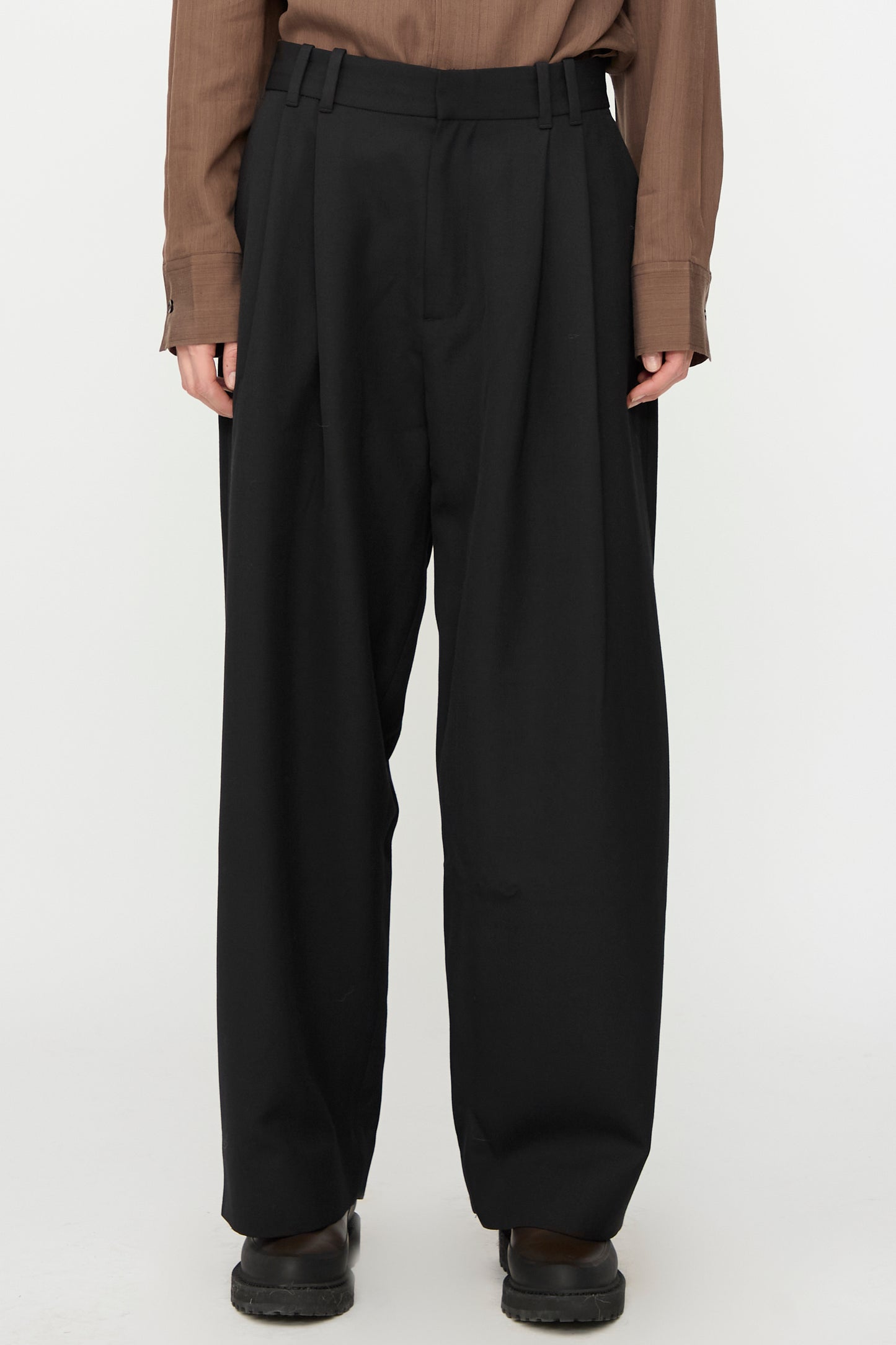 A person wears Studio Nicholson's Light Tech Wool Line Pant in Black, high-waisted and wide-leg with a tailored finish, paired with a brown long-sleeve shirt and black shoes, standing against a plain white background.