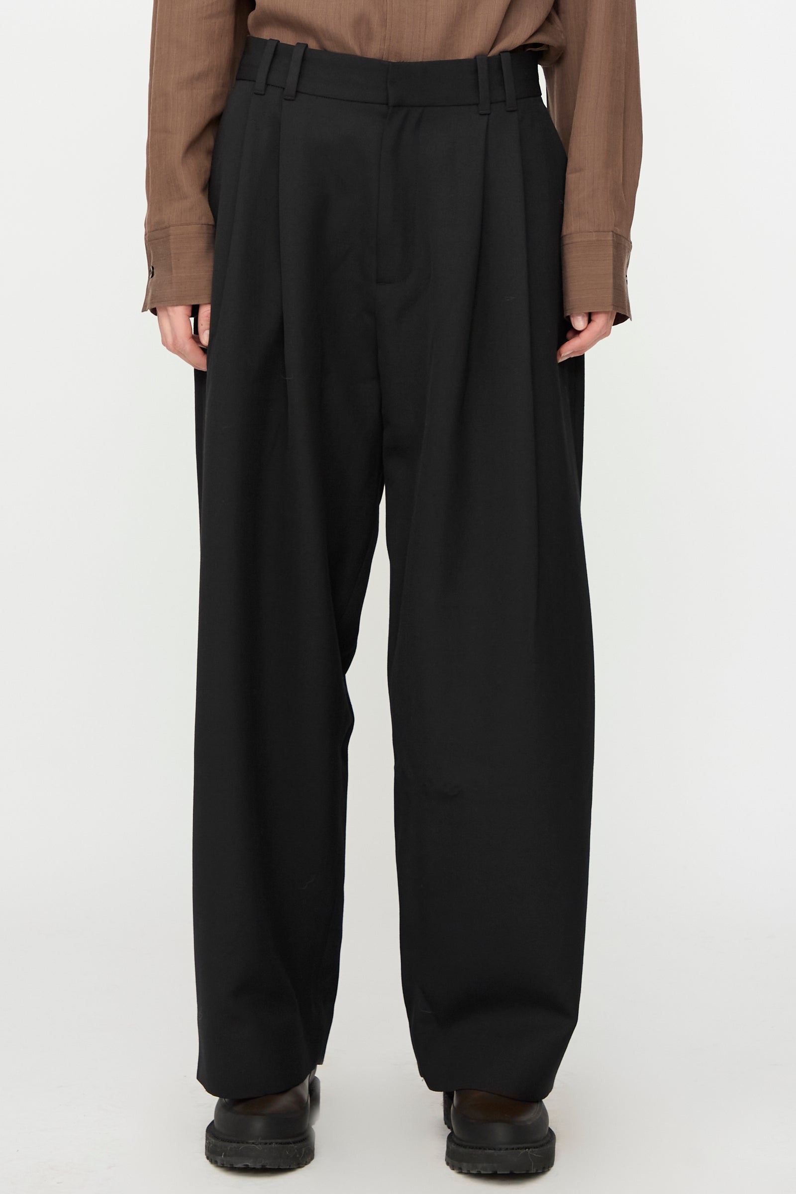 A person wears Studio Nicholson's Light Tech Wool Line Pant in Black, high-waisted and wide-leg with a tailored finish, paired with a brown long-sleeve shirt and black shoes, standing against a plain white background.