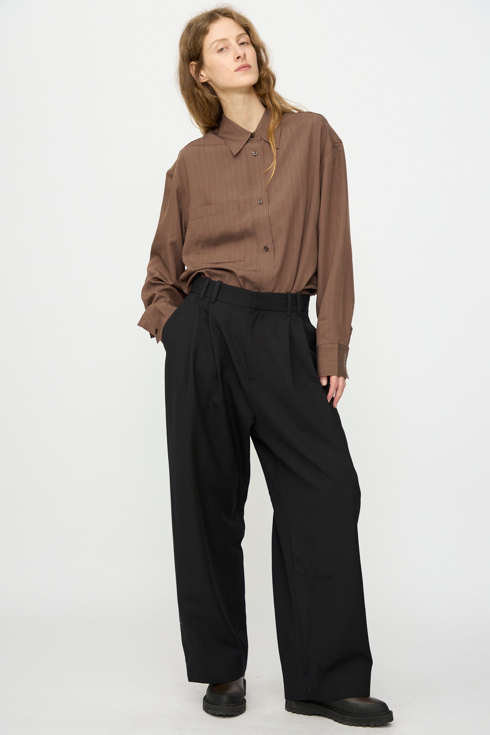 A person stands against a plain background wearing a brown button-up shirt, Studio Nicholson’s Light Tech Wool Line Pant in Black, and black shoes, with one hand in their pocket.