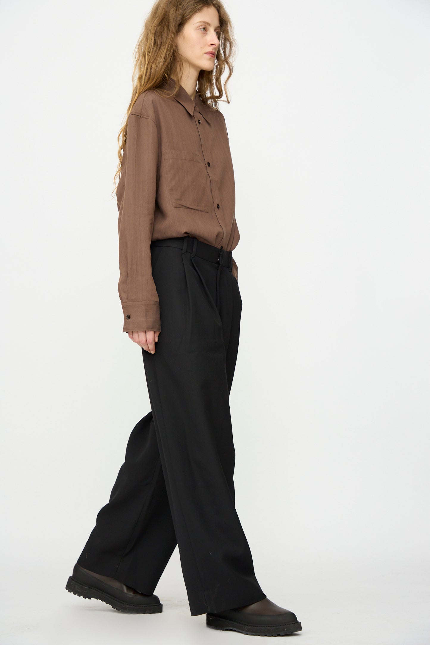 A person with long wavy hair stands against a white background, wearing a brown button-up shirt, black shoes, and Studio Nicholson's Light Tech Wool Line Pant in Black.