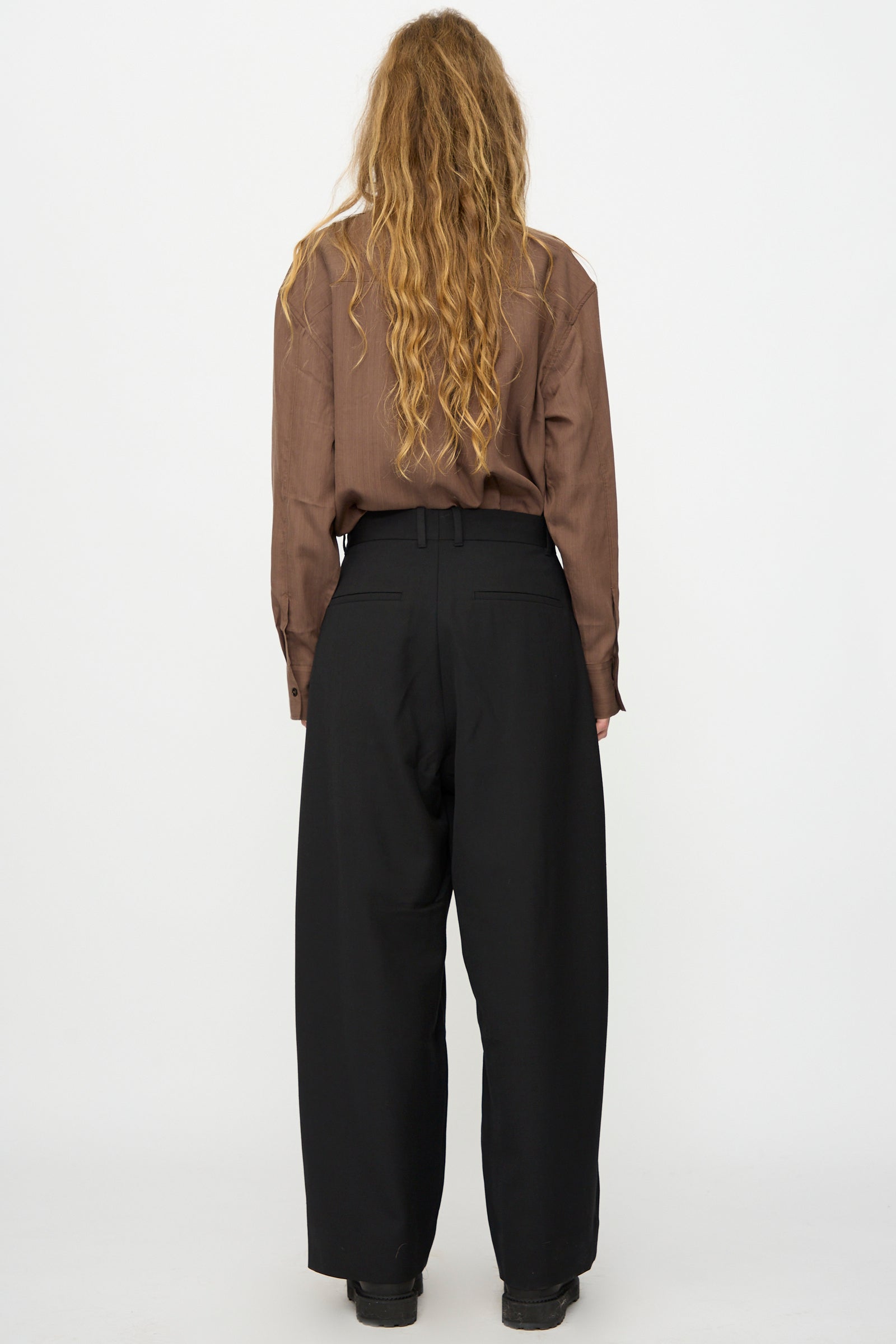Person with long wavy hair in a brown shirt and Studio Nicholson's Light Tech Wool Line Pant in Black, standing with back to camera against a plain white background, highlighting a relaxed yet tailored finish.