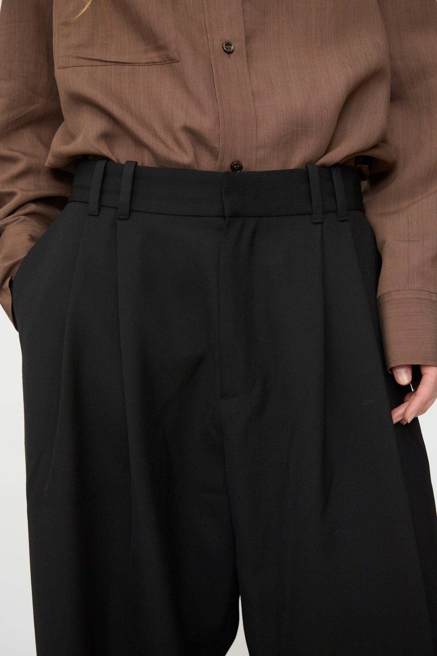 A person wears Studio Nicholson's Light Tech Wool Line Pant in Black—high-waisted, pleated, and tailored—with a brown button-up shirt tucked in and hands in pockets.