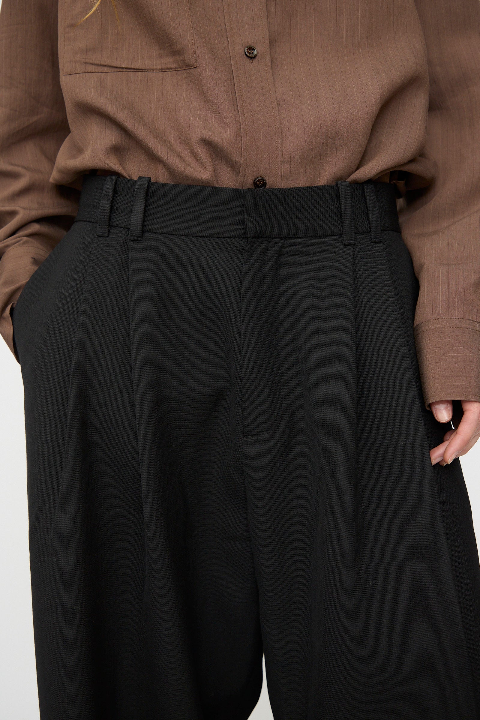A person wears Studio Nicholson's Light Tech Wool Line Pant in Black—high-waisted, pleated, and tailored—with a brown button-up shirt tucked in and hands in pockets.