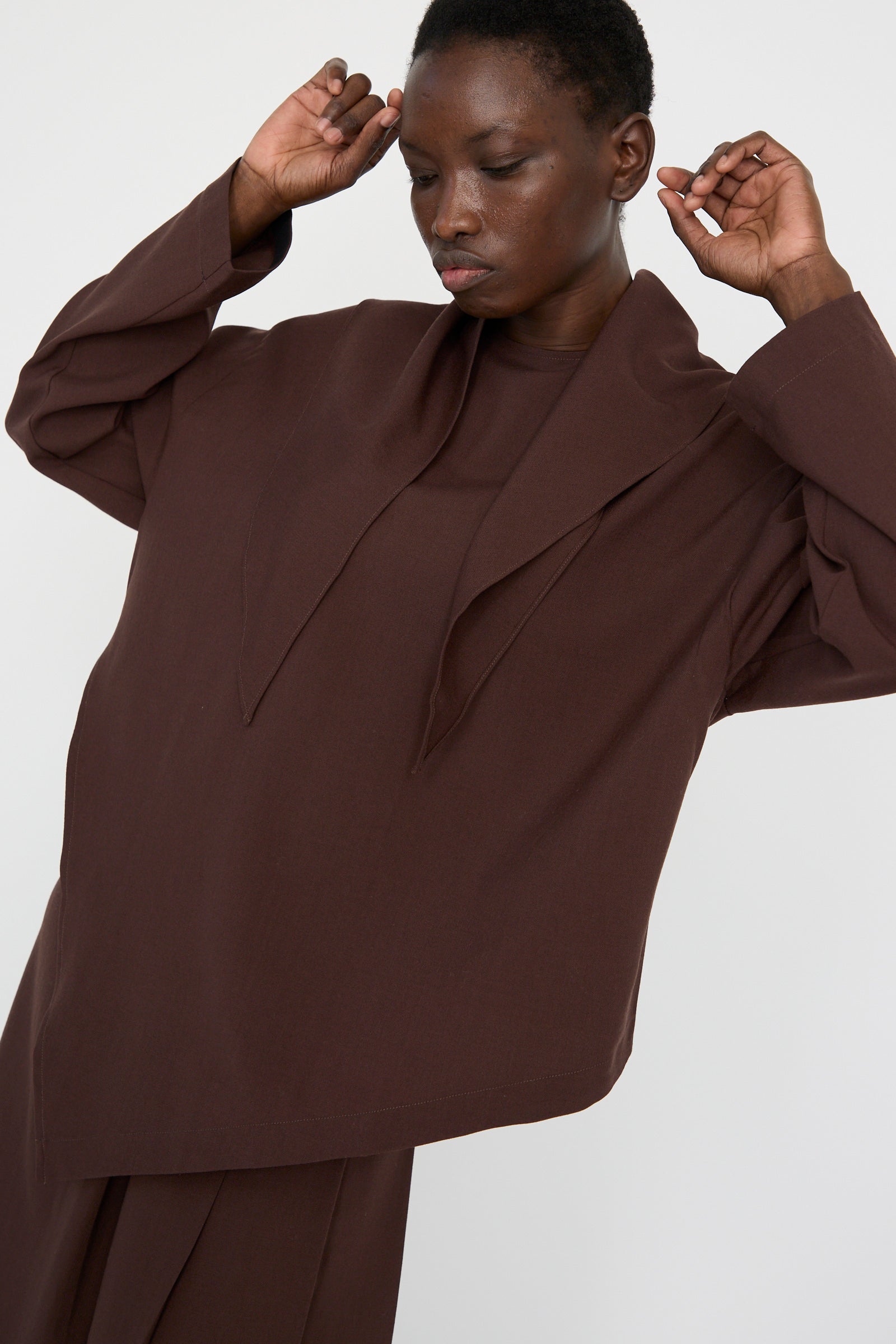 A model adjusts the detachable scarf of the Studio Nicholson Lightweight Textured Wool Bute Top in Conker, featuring an oversized design with a draped collar, against a plain background, with arms raised. 