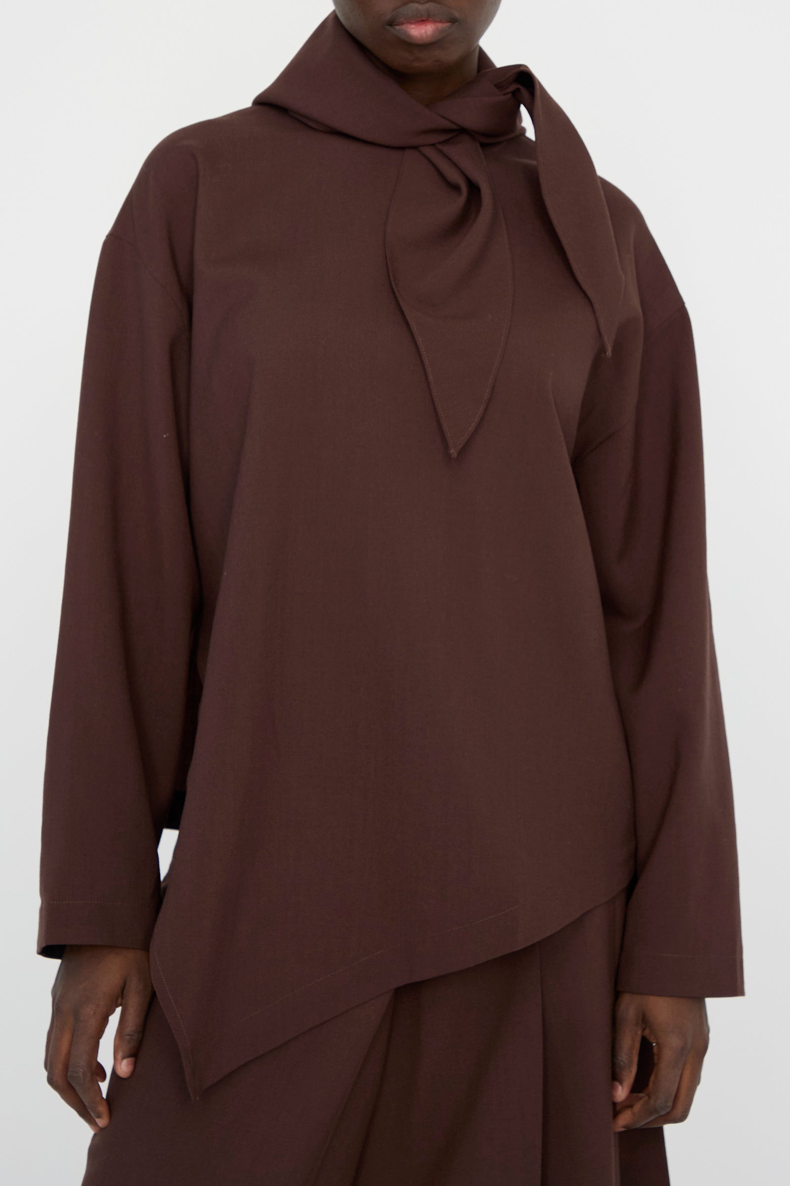 A person is wearing Studio Nicholson's "Lightweight Textured Wool Bute Top in Conker," a brown long-sleeve top with an asymmetrical hemline and a detachable scarf.