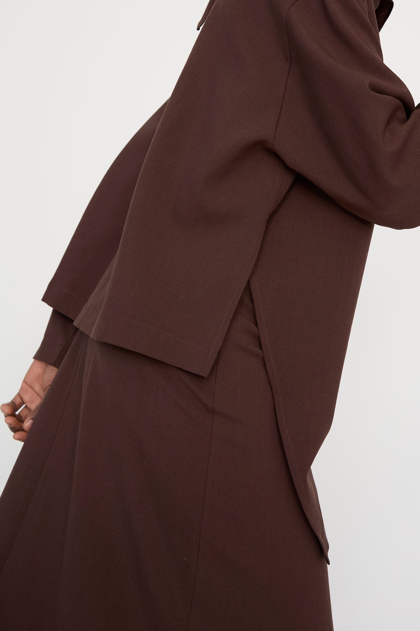 A model wears the Studio Nicholson Lightweight Textured Wool Bute Top in Conker, featuring long sleeves and an asymmetrical hemline, along with a matching skirt, against a plain backdrop.