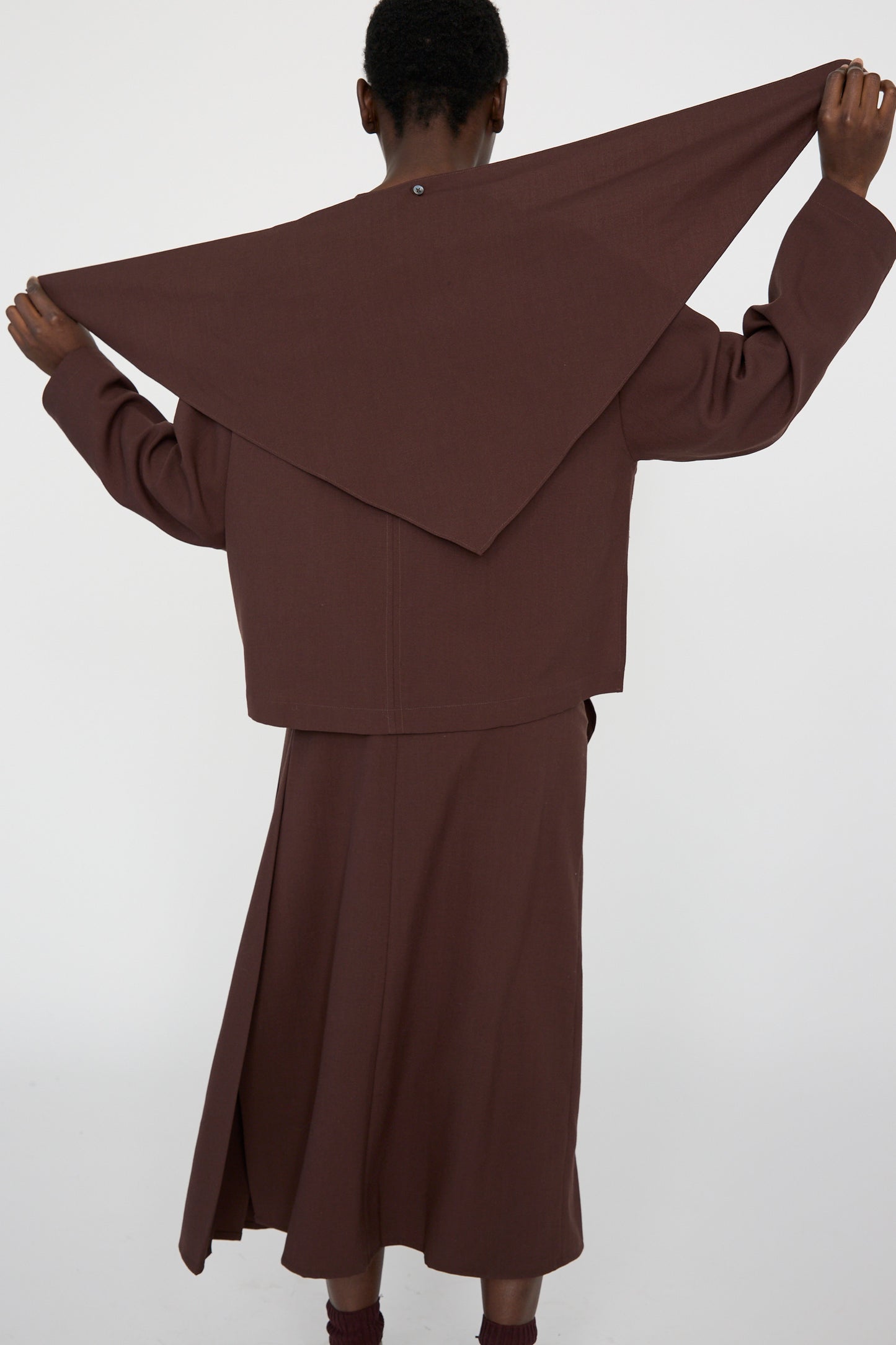 A person in a brown outfit with an asymmetrical hemline holds the Studio Nicholson Lightweight Textured Wool Bute Top in Conker over their shoulders, standing against a plain white background, stretching the collar in a triangular shape. 