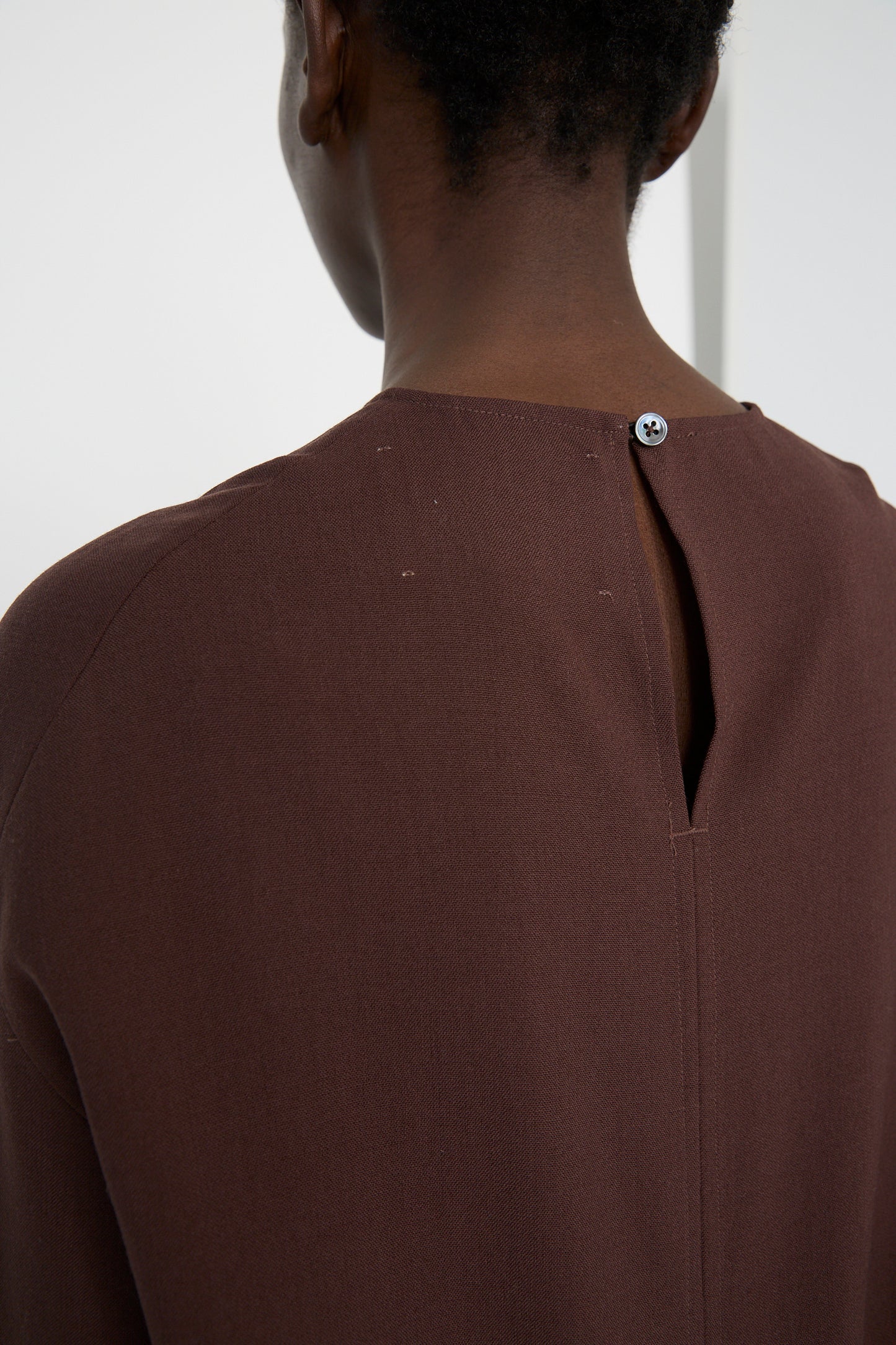 A person is seen from behind, wearing the Lightweight Textured Wool Bute Top in Conker by Studio Nicholson, featuring a buttoned neckline and an asymmetrical hemline.