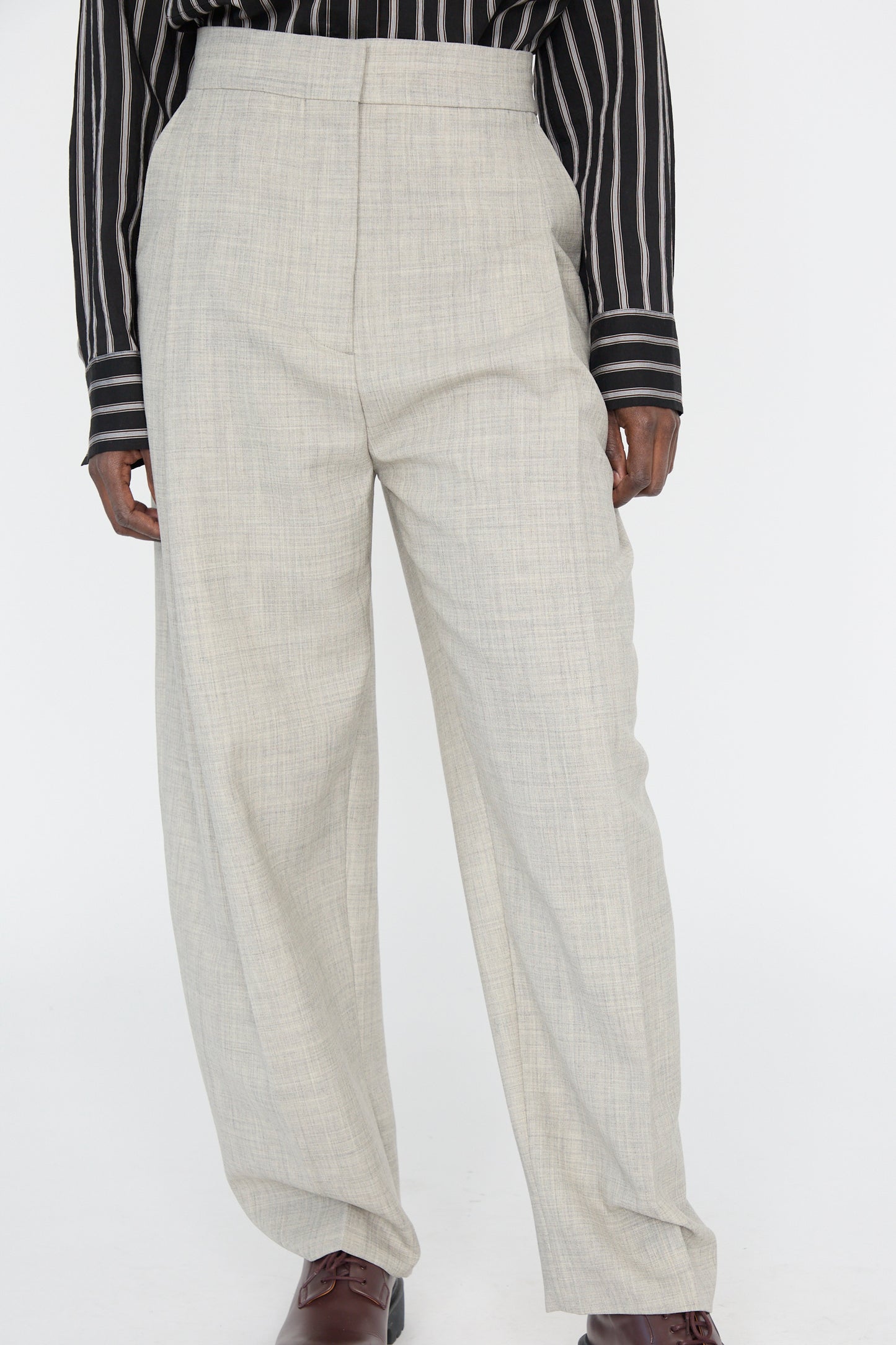 A person stands against a white background wearing Studio Nicholson's Lightweight Tropical Wool Vai Trouser in Ice Melange, featuring a tailored fit and high waistband, paired with a black and white striped shirt.