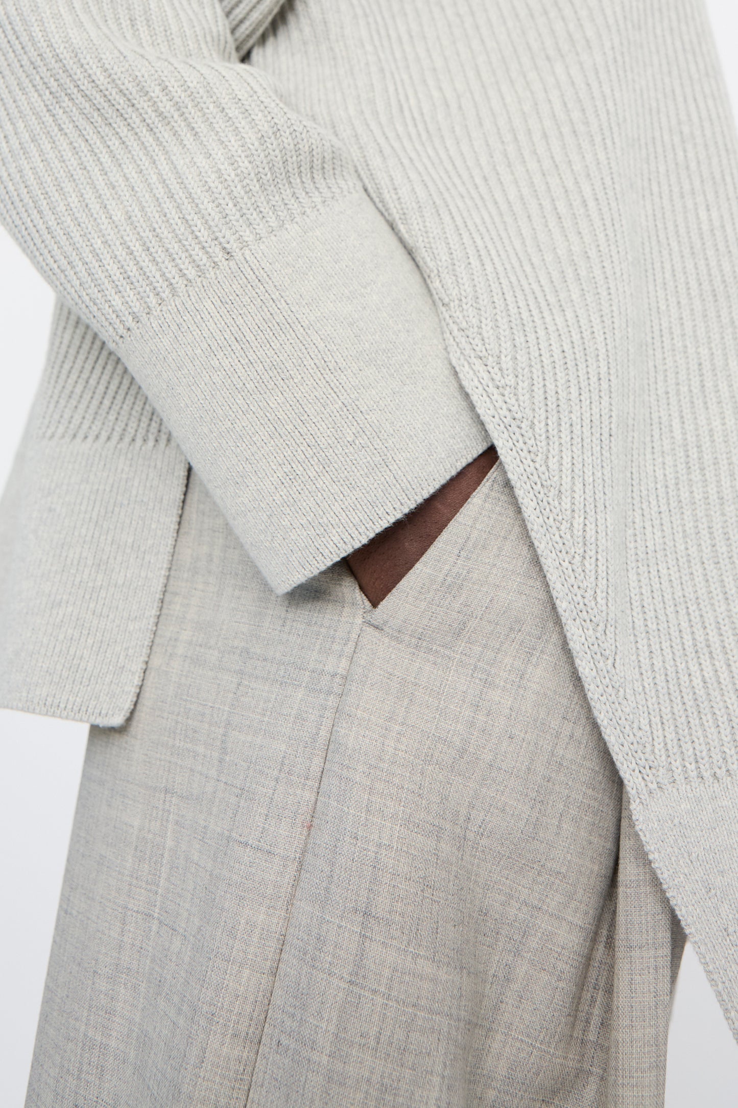 A close-up shot of a person wearing a light gray ribbed sweater and Studio Nicholson's Lightweight Tropical Wool Vai Trouser in Ice Melange has one hand in their pocket.