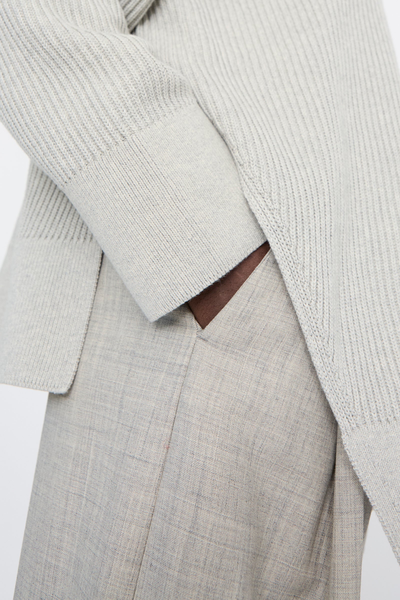 A close-up shot of a person wearing a light gray ribbed sweater and Studio Nicholson's Lightweight Tropical Wool Vai Trouser in Ice Melange has one hand in their pocket.