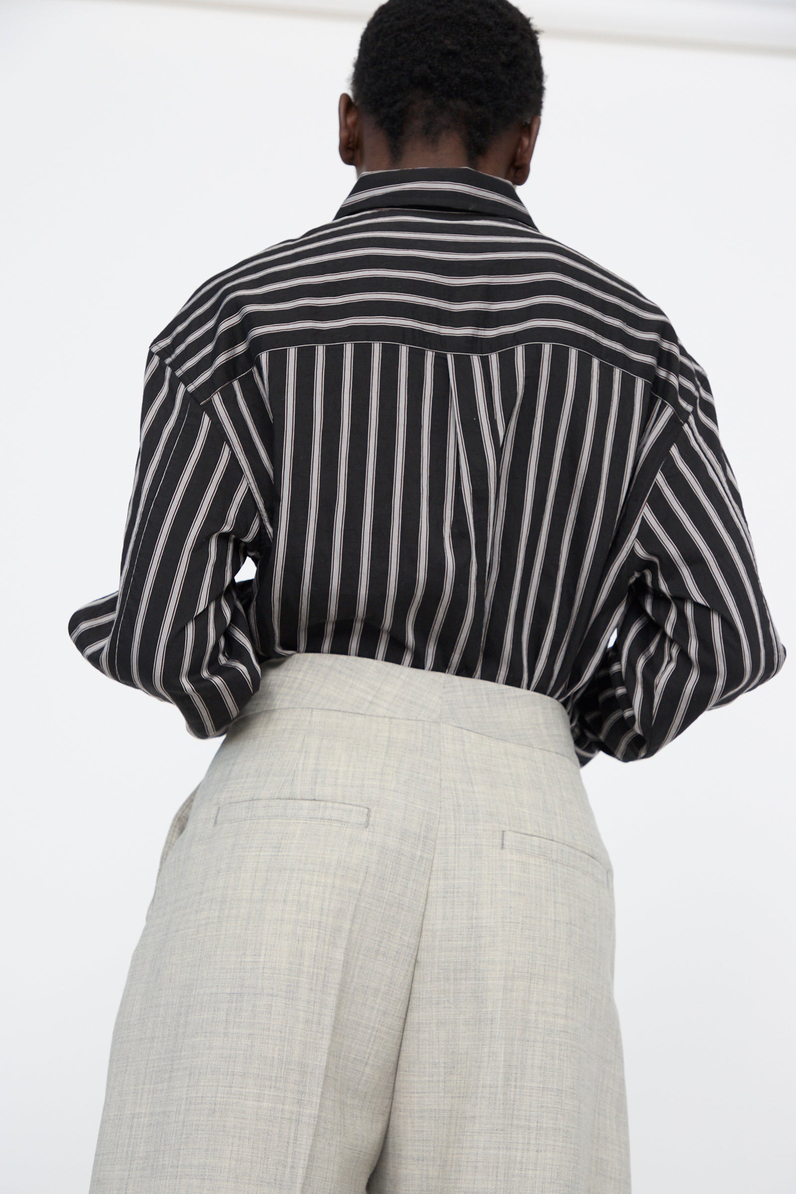 A person is seen from behind wearing a striped dark shirt and Studio Nicholson's Lightweight Tropical Wool Vai Trouser in Ice Melange, featuring tailored design and a high waistband.