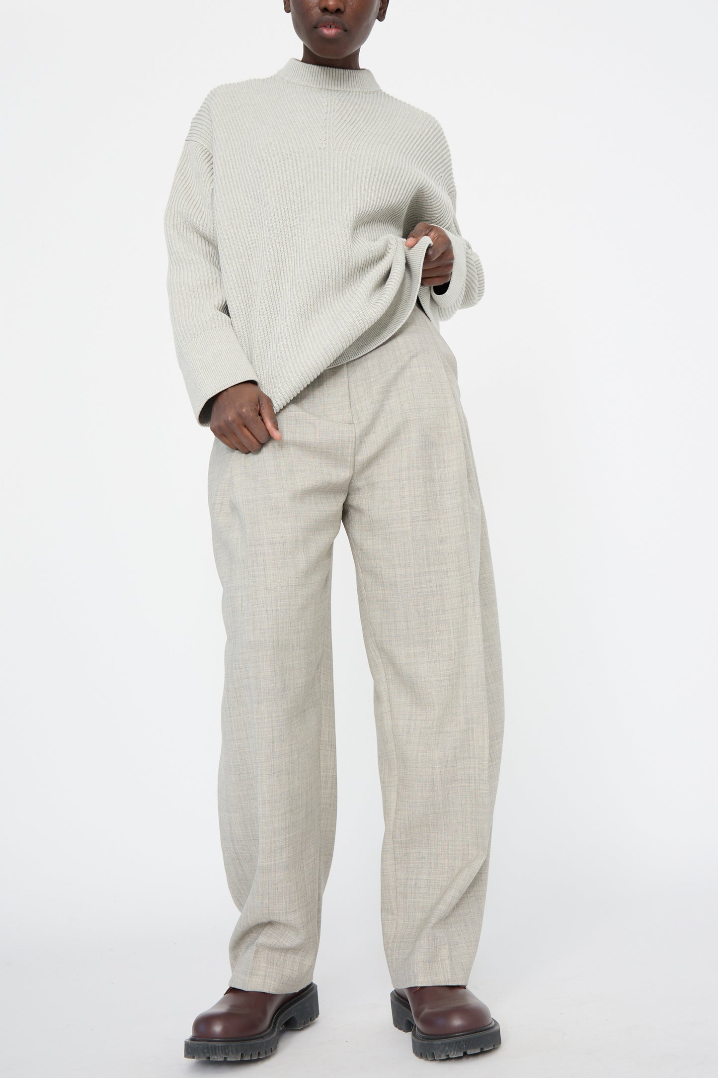 A person stands against a white background, wearing Studio Nicholson's Lightweight Tropical Wool Vai Trouser in Ice Melange and a light gray knit sweater, lifting the bottom hem.