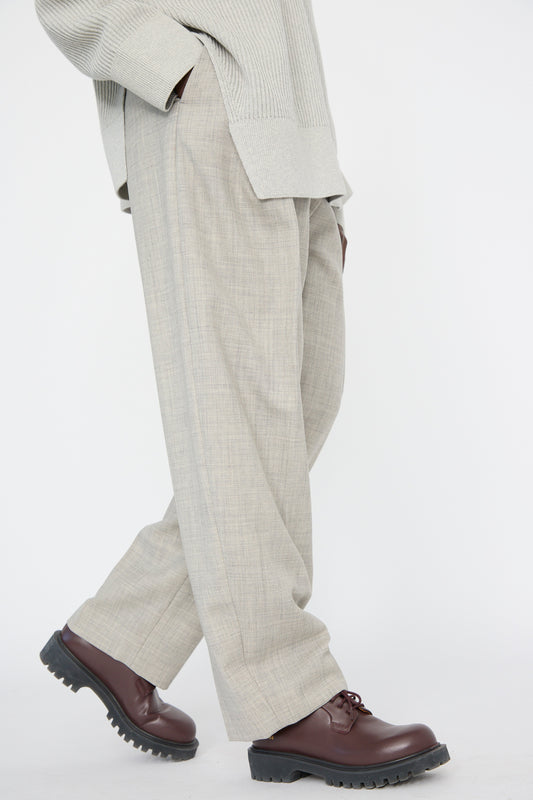 A person wearing Studio Nicholson's Lightweight Tropical Wool Vai Trouser in Ice Melange, paired with a light gray sweater and brown dress shoes, stands against a plain background.