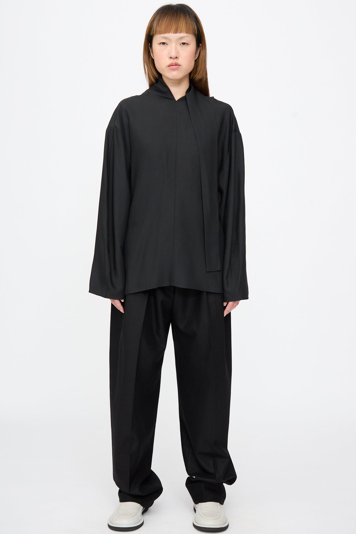 A person wearing a relaxed-fit, black Loti Draped Tie Neck Top in black viscose wool with a draped tie neck and matching black trousers.