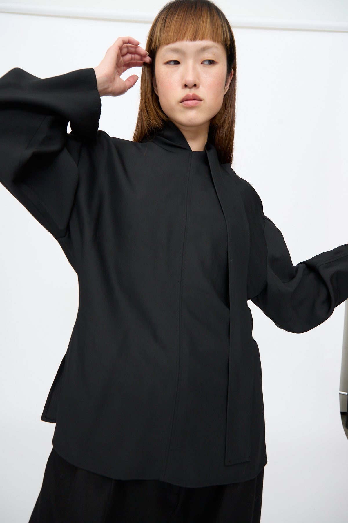 A model wearing the black Studio Nicholson Loti Draped Tie Neck Top, featuring a relaxed-fit, long-sleeved silhouette with a high neckline and vertical front tie detail.