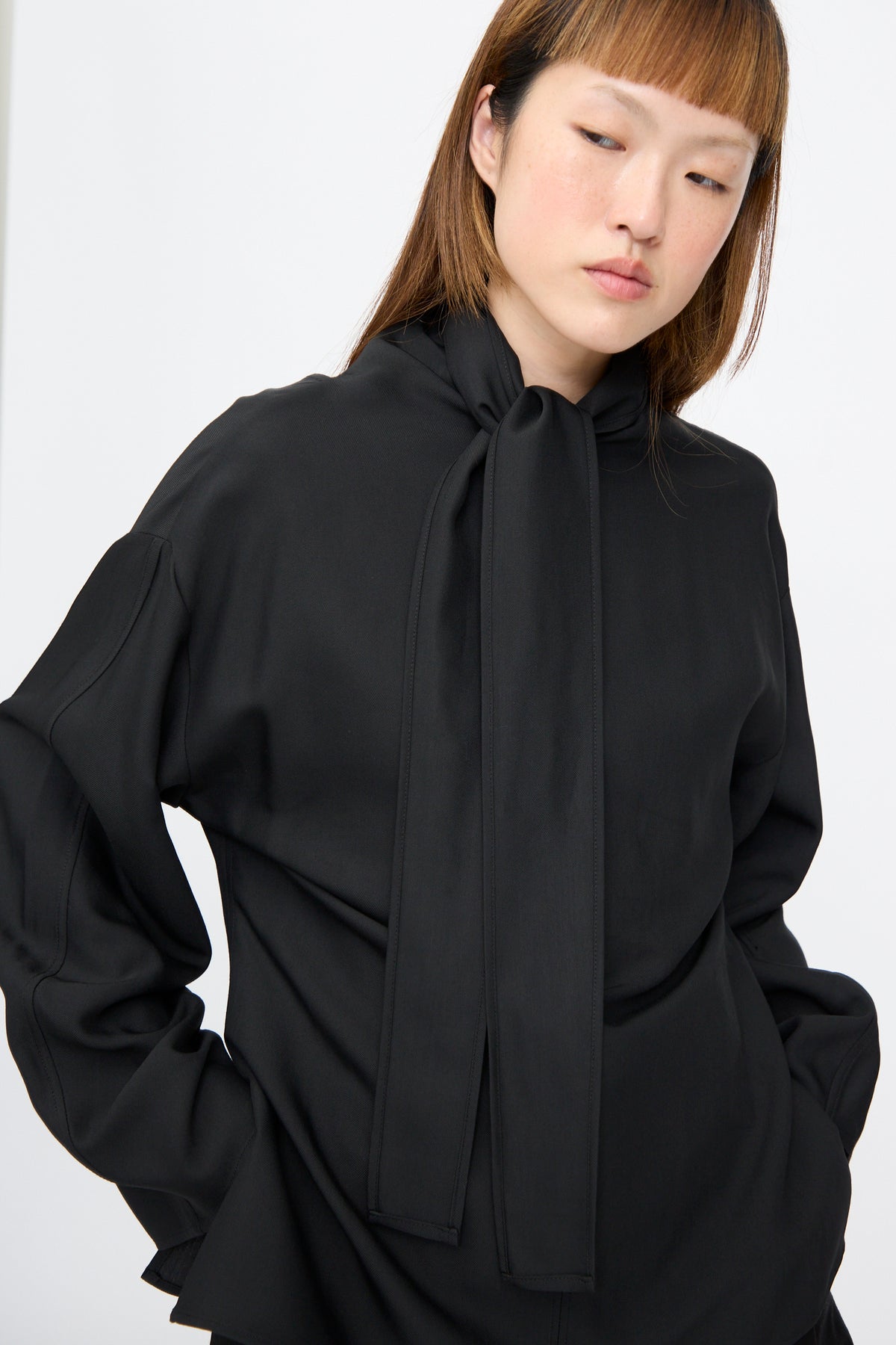 A person wearing a black Studio Nicholson Loti Draped Tie Neck Top, a panel-cut viscose wool piece with a draped tie-neck detail.