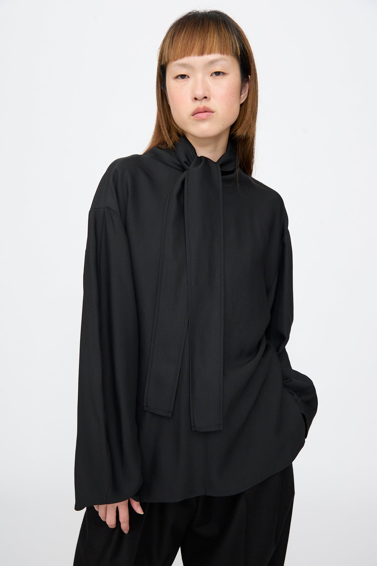A person wearing the Studio Nicholson Loti Draped Tie Neck Top in black, featuring a relaxed fit, long sleeves, and a draped tie at the neck.