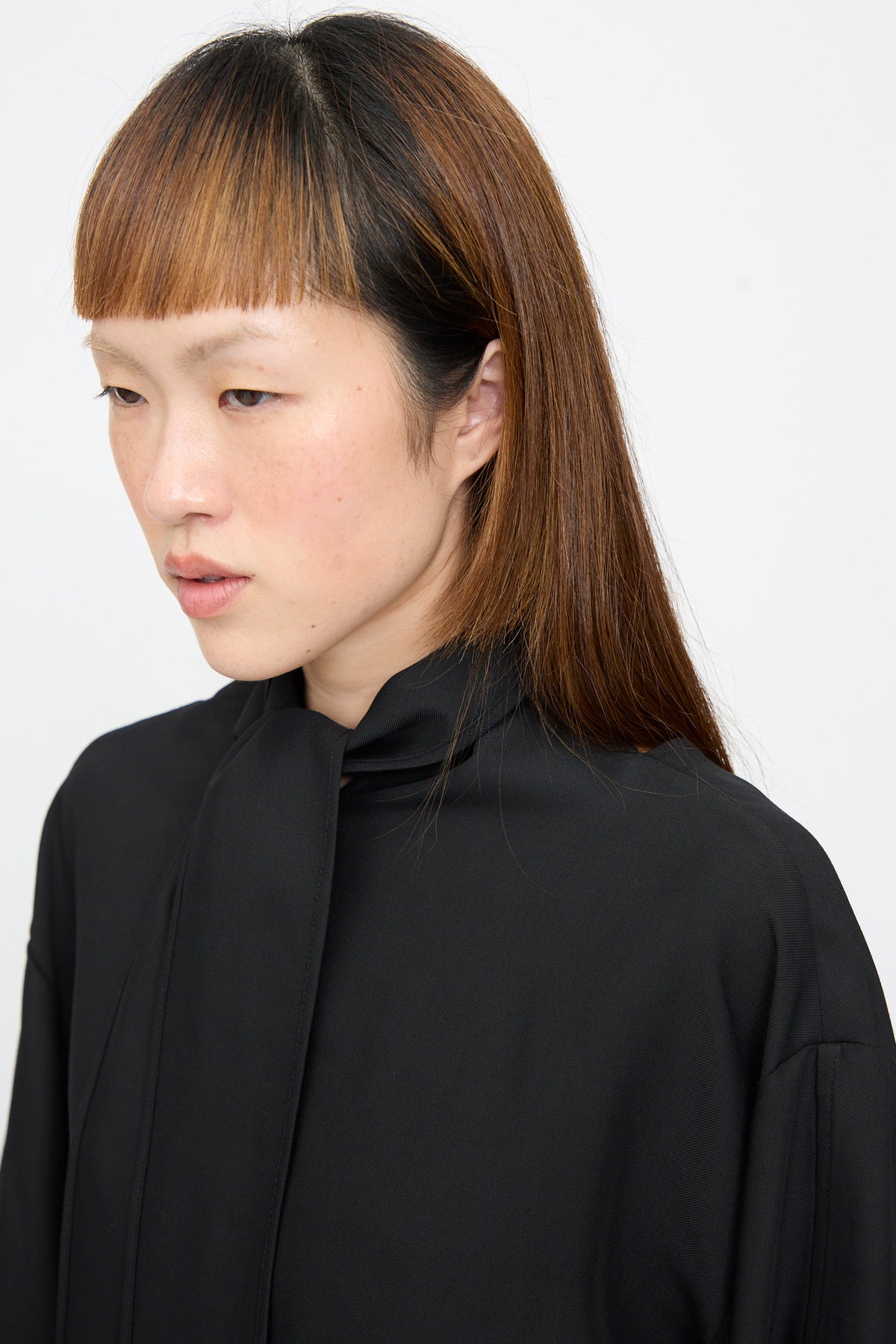 A person wears a black Studio Nicholson Loti Draped Tie Neck Top with a relaxed fit and a draped tie at the neckline.