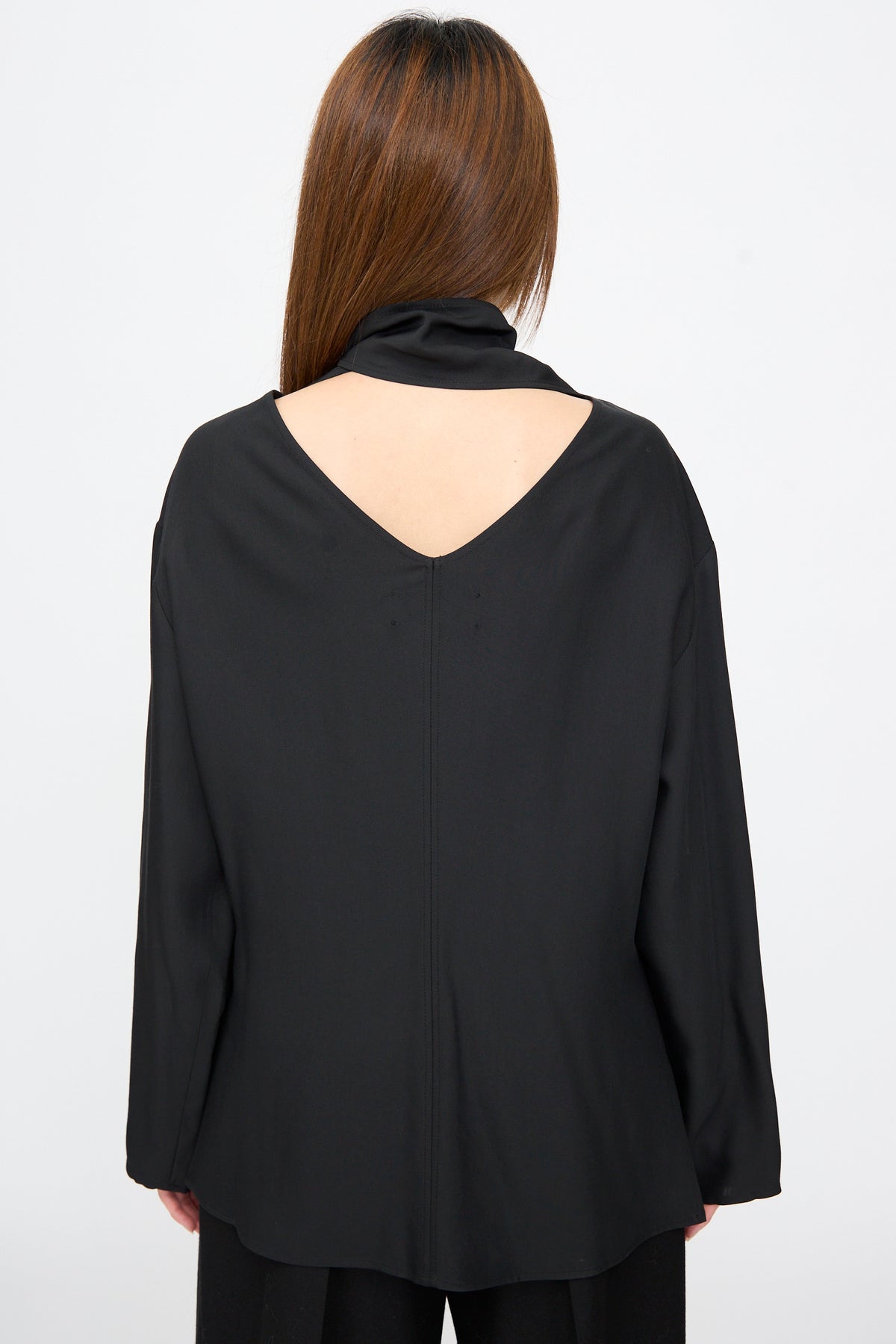 Back view of the Studio Nicholson Loti Draped Tie Neck Top in black, a relaxed-fit, long-sleeved panel-cut shirt with a V-neckline and high draped collar.