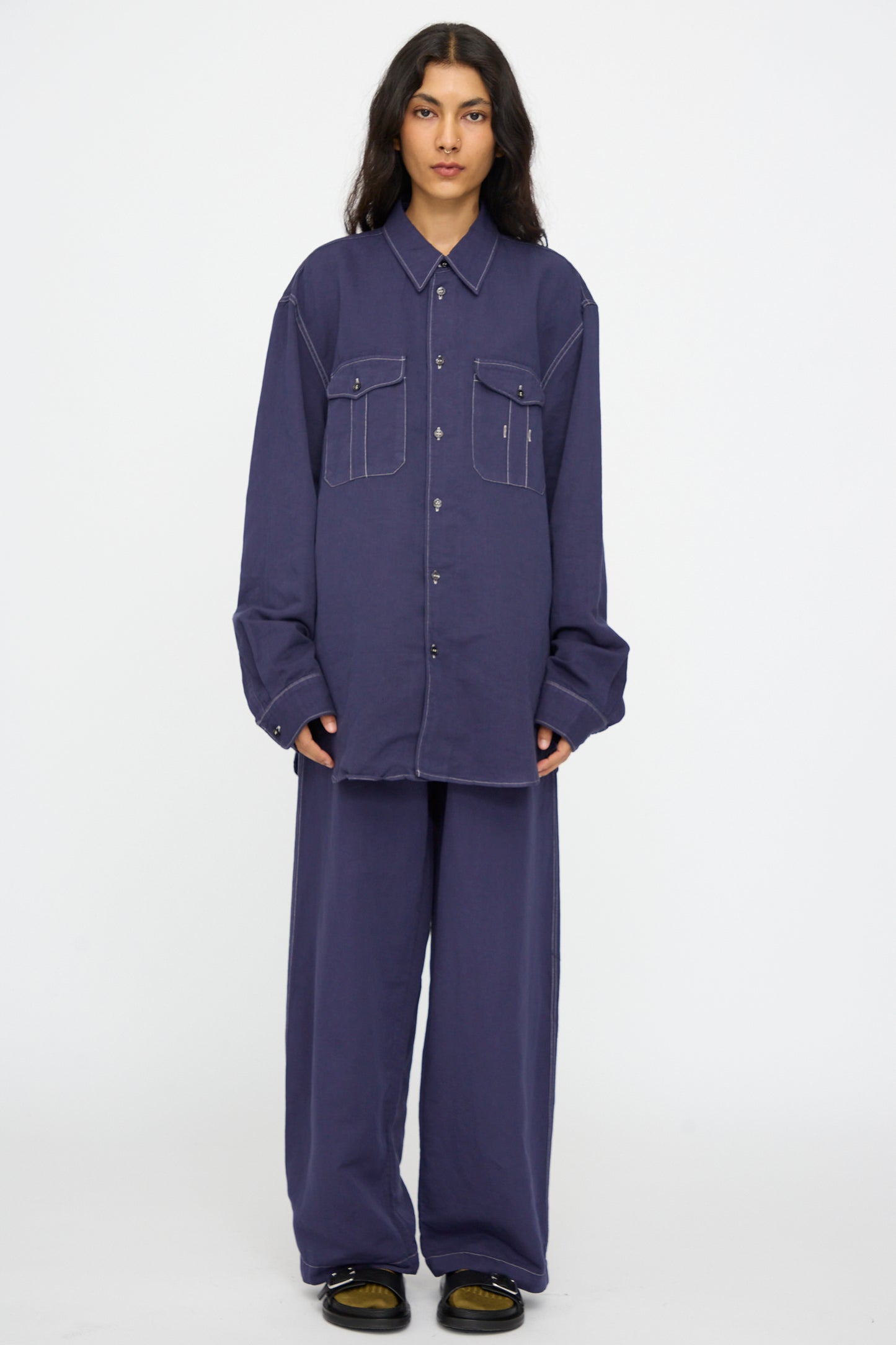 A person wears the Studio Nicholson Louro Utility Pocket Shirt in Indigo, a relaxed-fit, long-sleeve linen top with two front pockets, paired with matching wide-leg pants, standing against a plain white background.