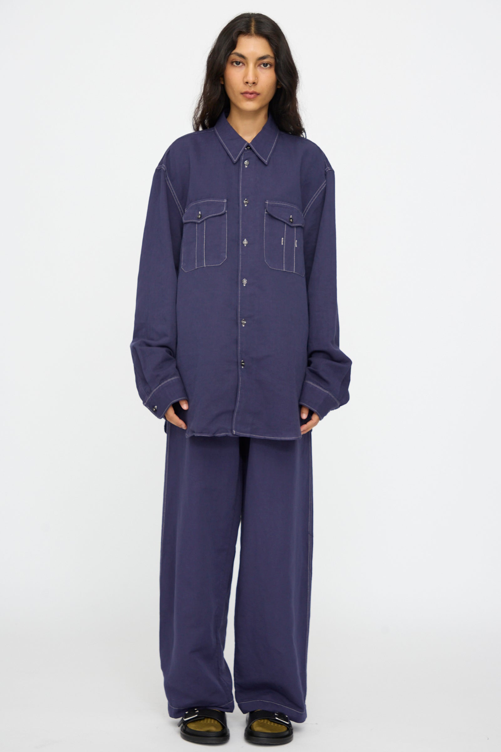 A person wears the Studio Nicholson Louro Utility Pocket Shirt in Indigo, a relaxed-fit, long-sleeve linen top with two front pockets, paired with matching wide-leg pants, standing against a plain white background.
