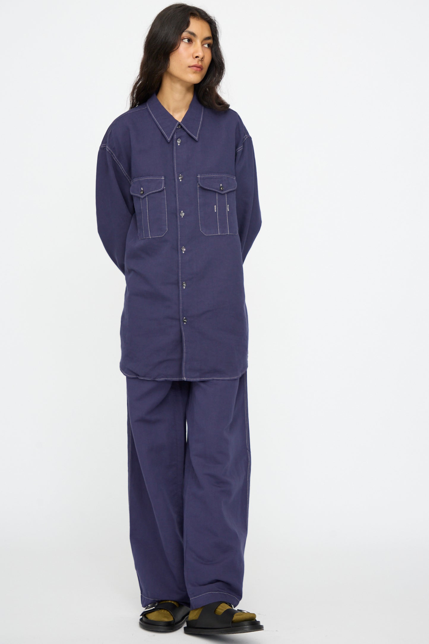 A person stands against a plain background wearing the Studio Nicholson Louro Utility Pocket Shirt in Indigo, featuring a relaxed fit and large front pockets, paired with wide-leg pants and black and olive green shoes.