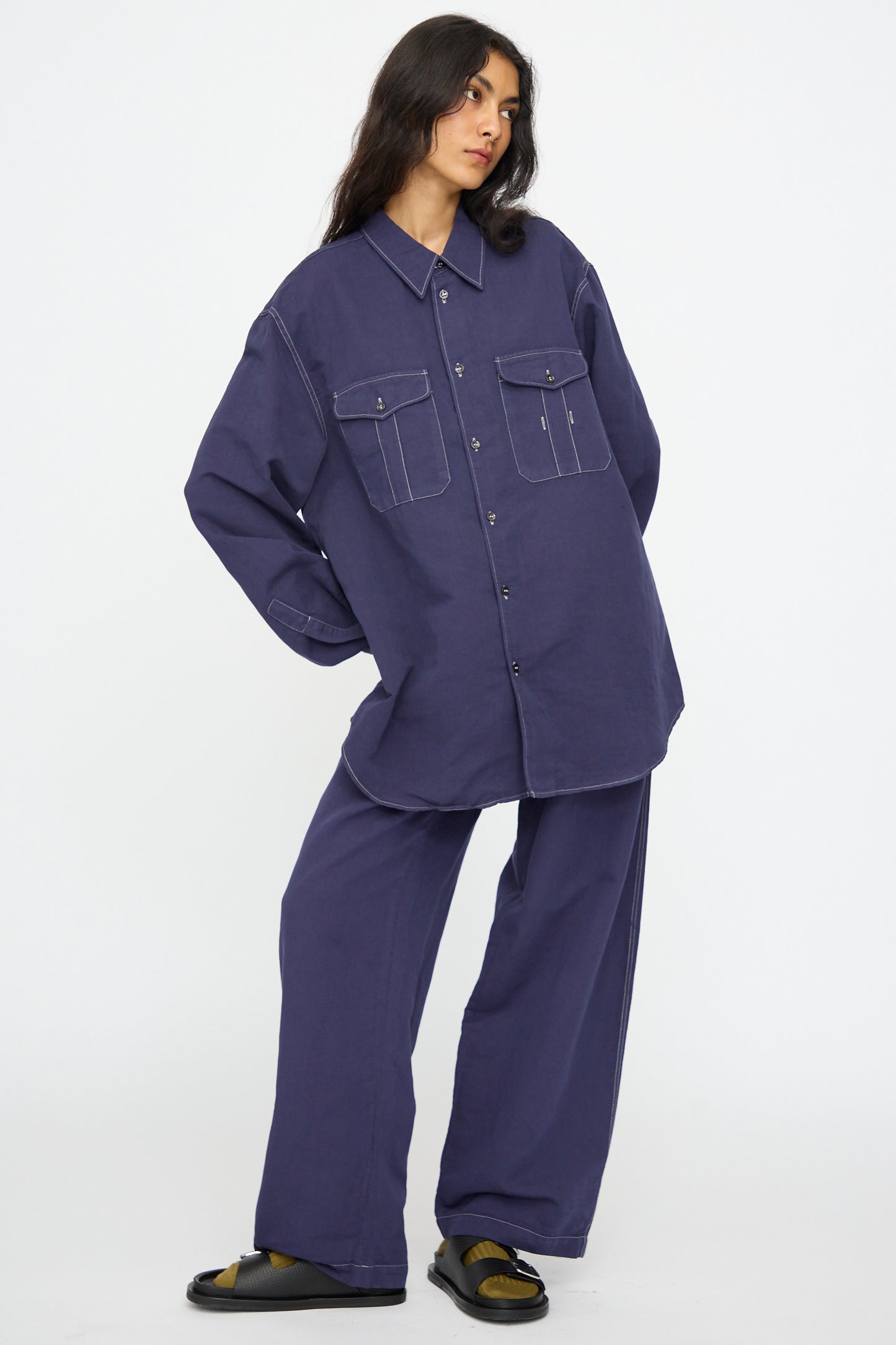 A person wears the Studio Nicholson Louro Utility Pocket Shirt in Indigo with large front pockets and matching relaxed fit pants, standing against a plain white background.