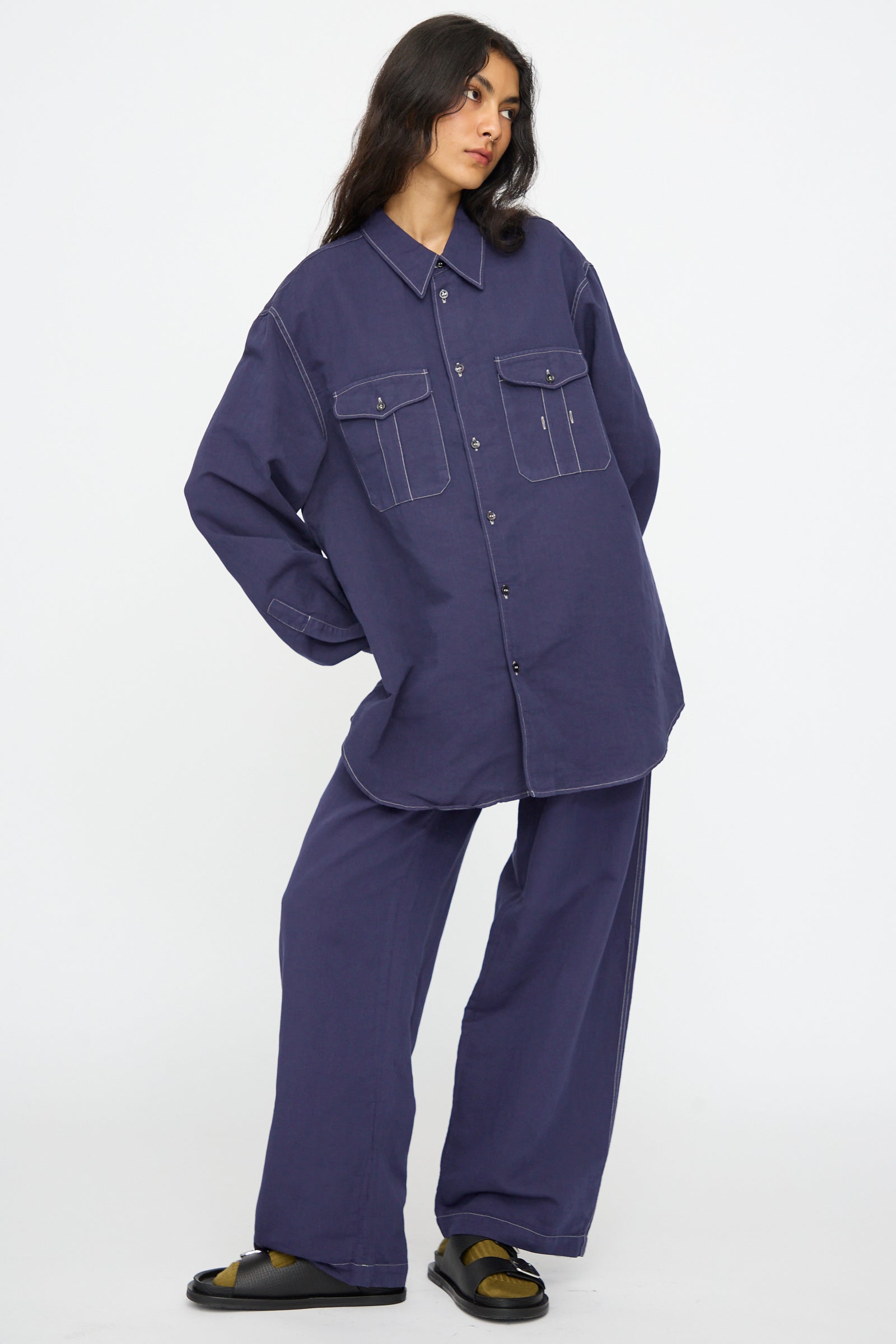 A person wears the Studio Nicholson Louro Utility Pocket Shirt in Indigo with large front pockets and matching relaxed fit pants, standing against a plain white background.