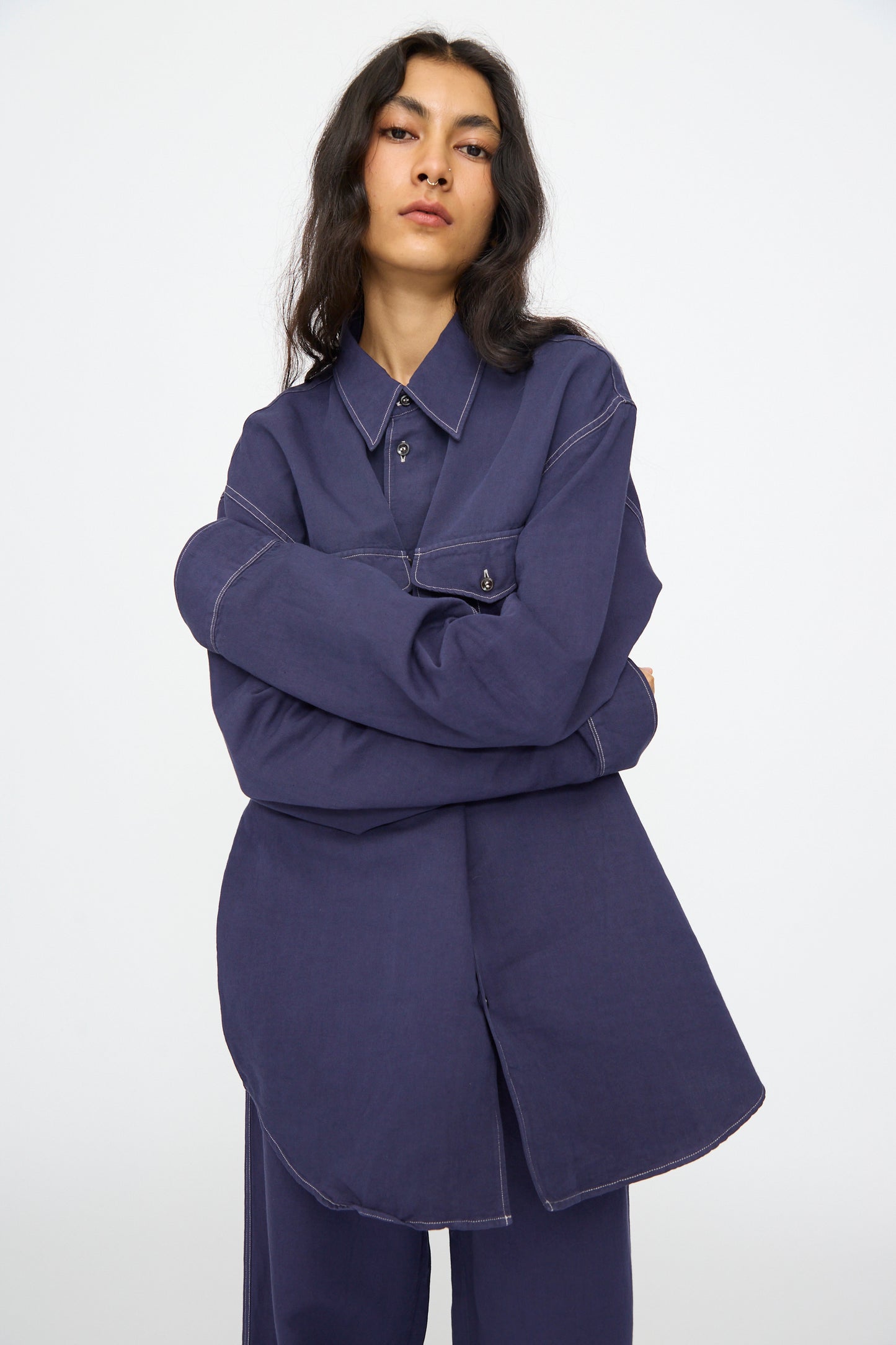 A person with long dark hair wears the Studio Nicholson Louro Utility Pocket Shirt in Indigo, styled with matching pants, standing against a plain light background.
