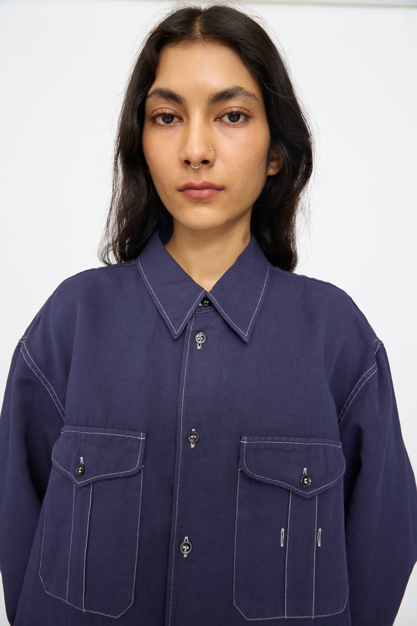 A person with long dark hair wears the Studio Nicholson Louro Utility Pocket Shirt in indigo, featuring two front pockets, standing against a plain white background.