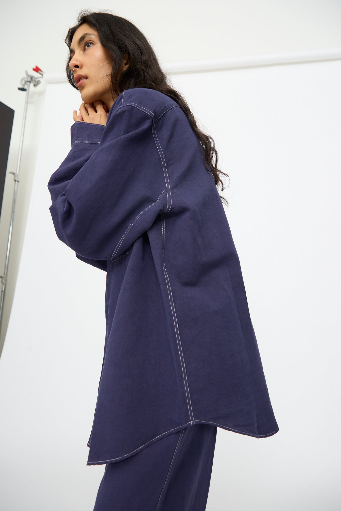 A woman models the Studio Nicholson Louro Utility Pocket Shirt in Indigo, styled with matching pants. She stands against a white backdrop, gazing sideways with her hands near her face.